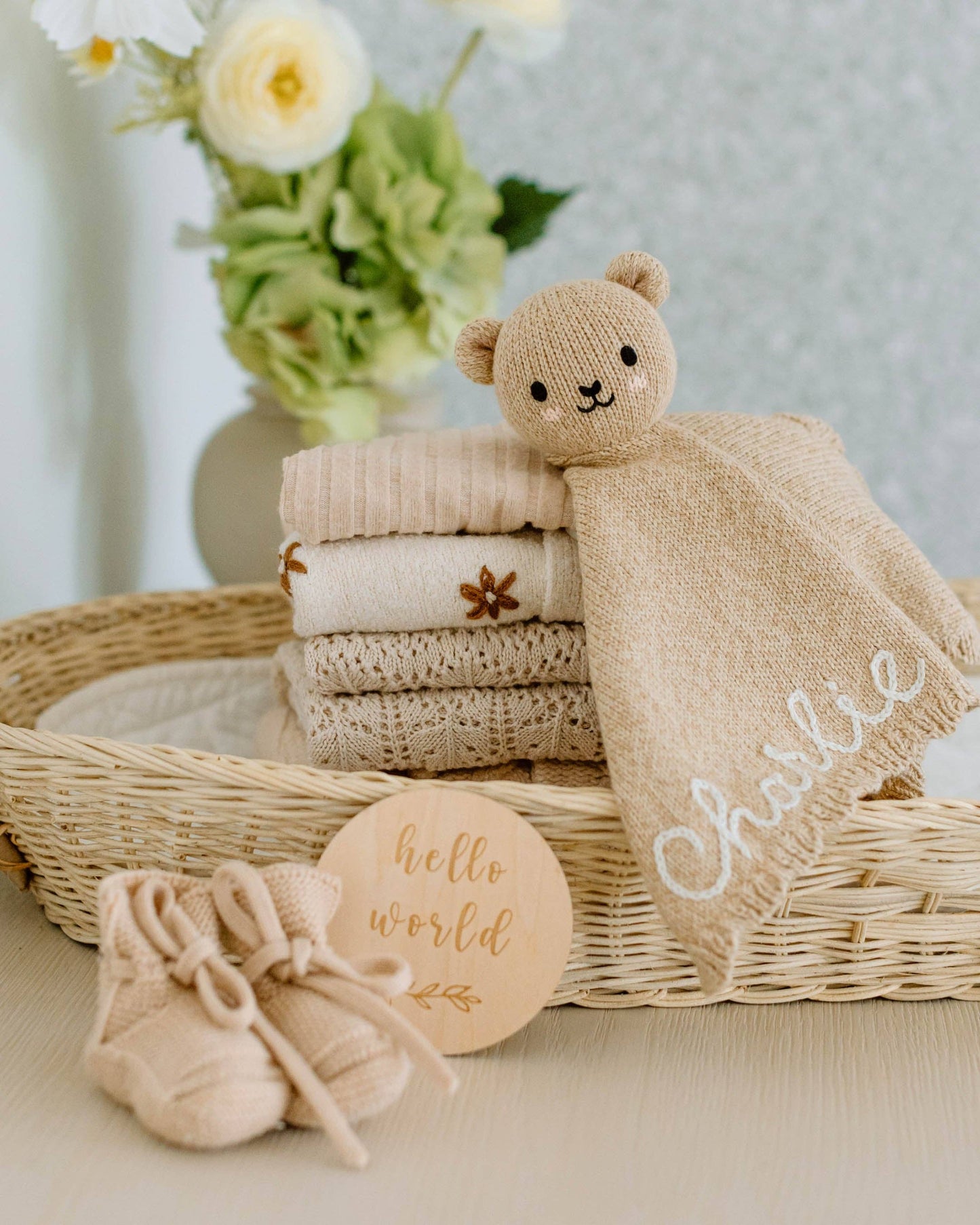 Baby items including a teddy bear lovey, folded blankets, and baby shoes in a basket with a 'hello world' sign.