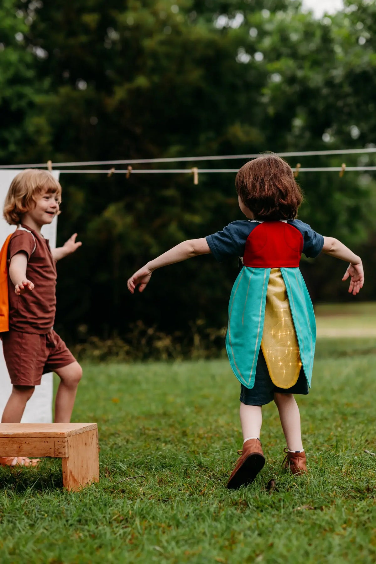Two children in colorful costumes playing outdoors on a grassy area with trees in the background.