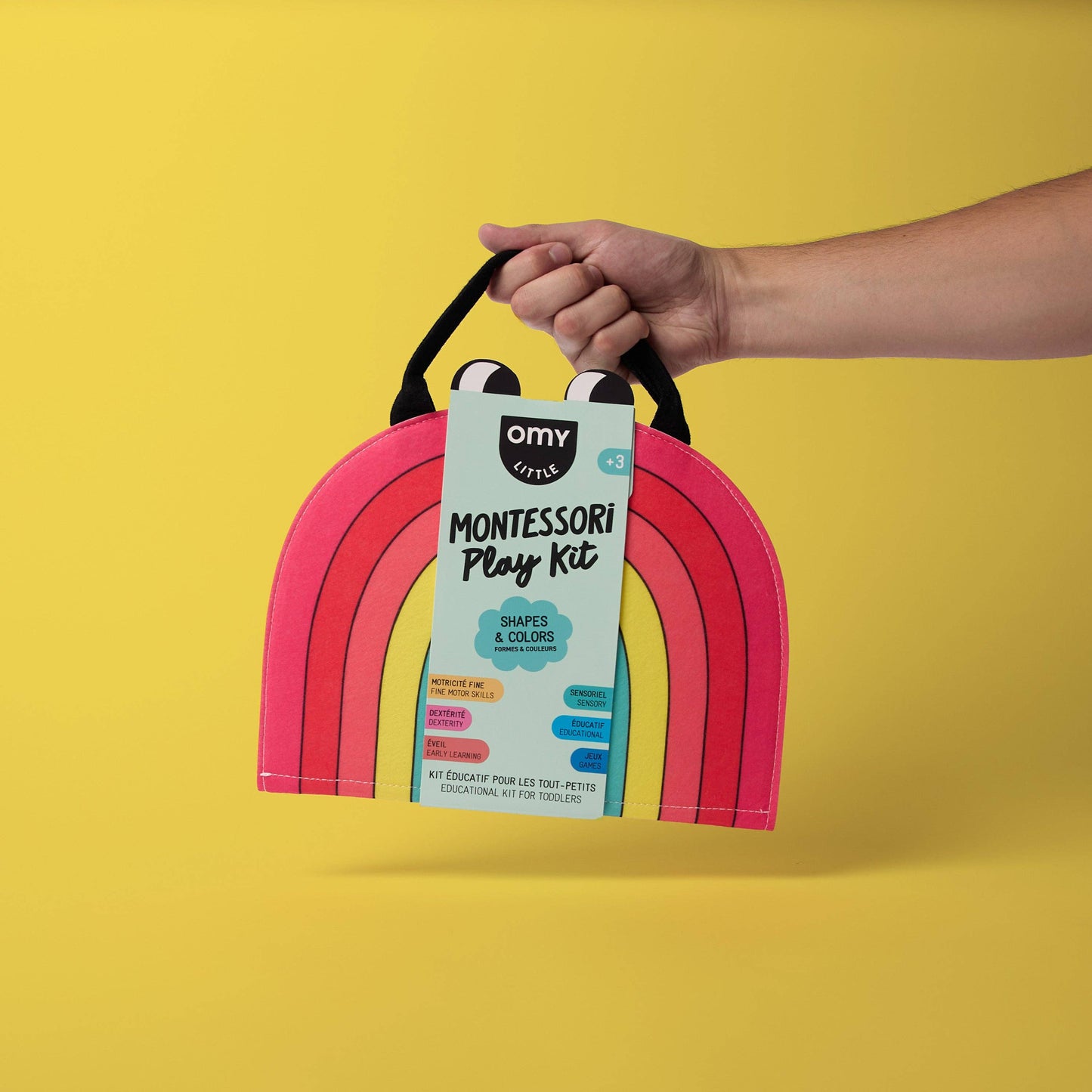 Hand holding a pink arch-shaped toy with a Montessori play kit label against a yellow background