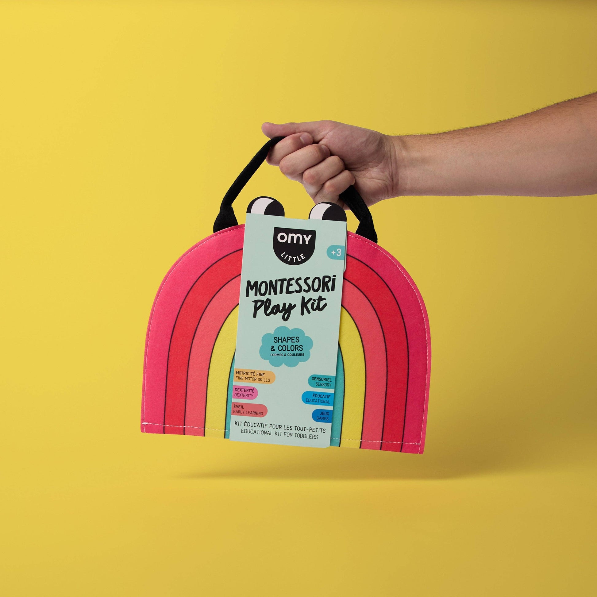 Hand holding a pink arch-shaped toy with a Montessori play kit label against a yellow background