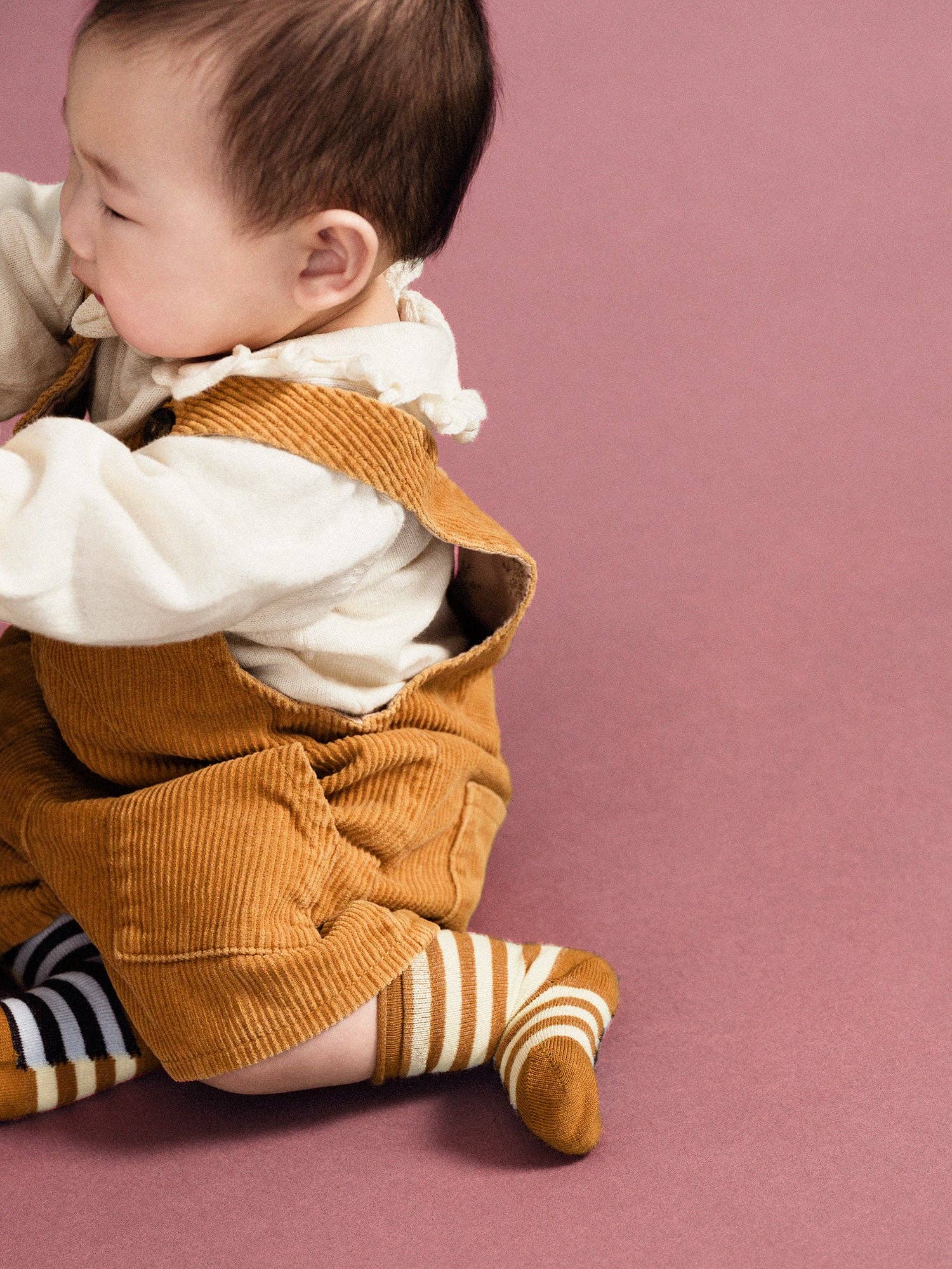Baby wearing a mustard yellow outfit with a white shirt and striped socks on a pink background
