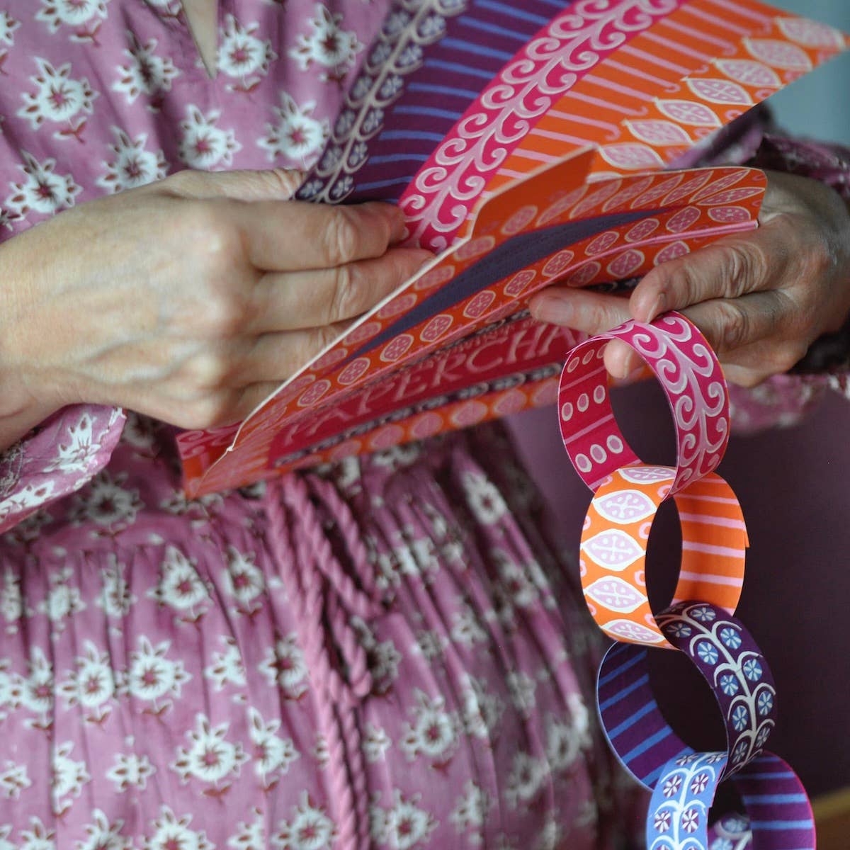 Hands separating patterned paper strips beside an assembled chain of pink, orange and purple links.