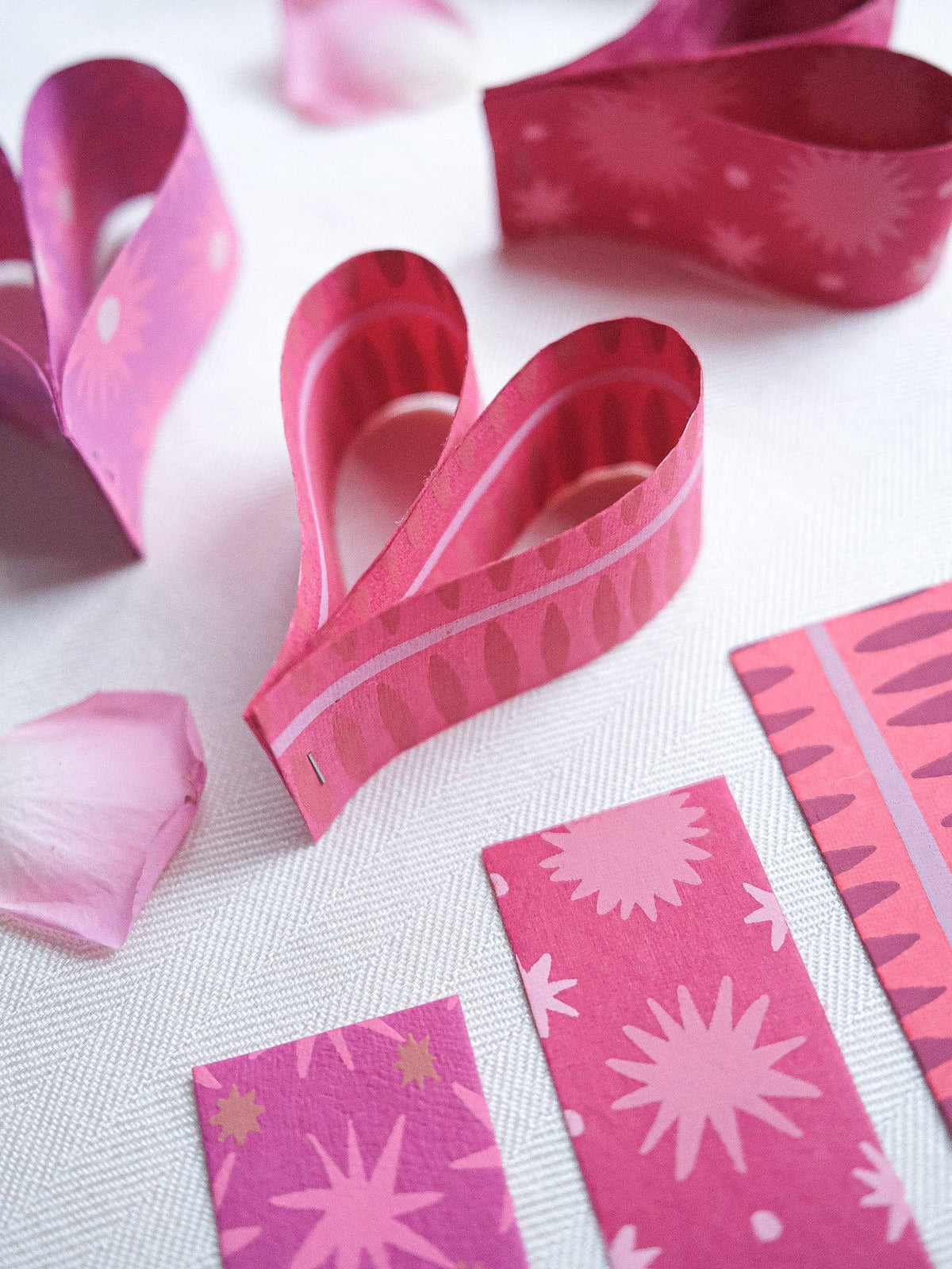 Close-up of pink patterned paper strips folded into hearts beside rose petals on white fabric.
