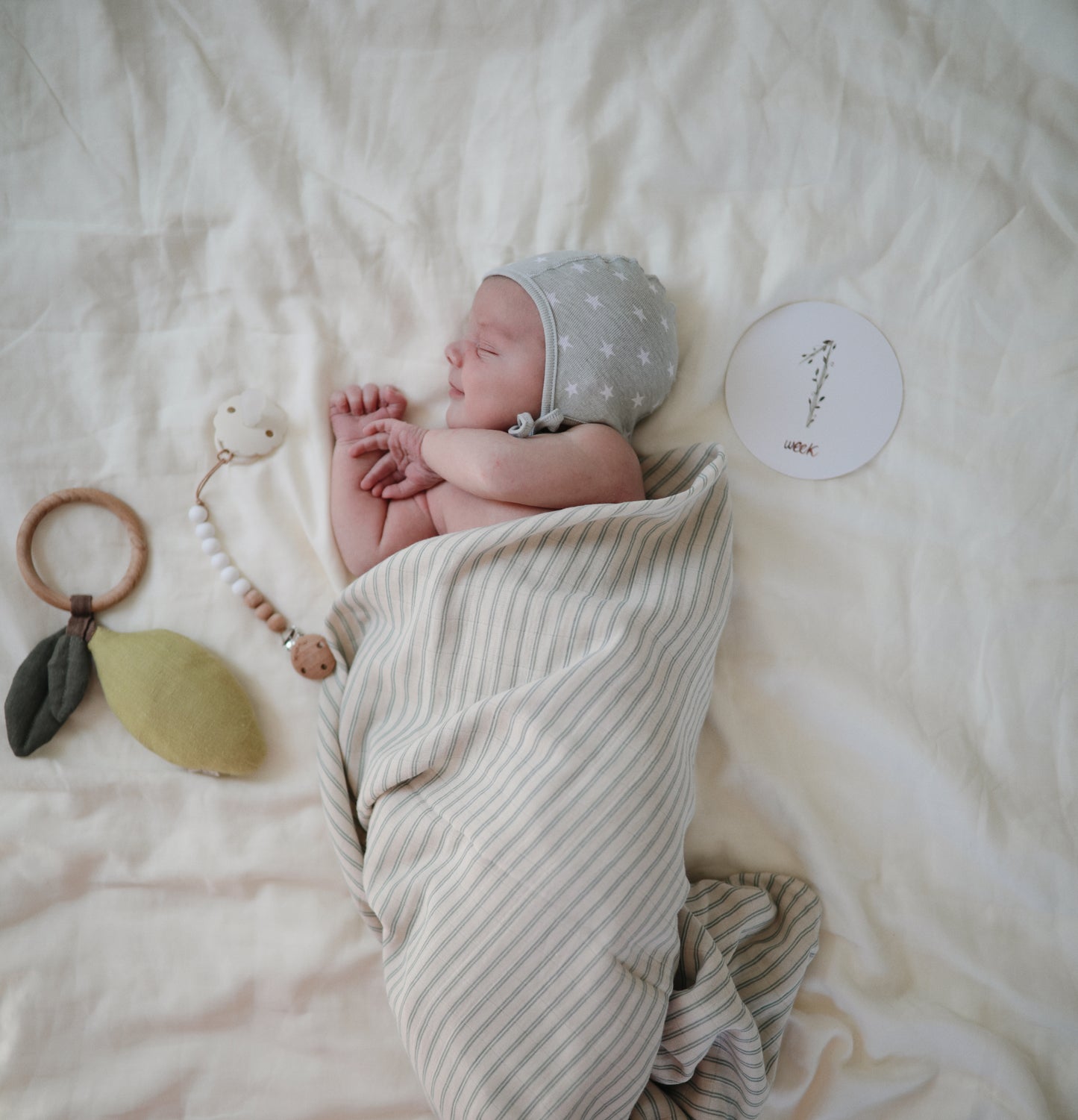 Newborn baby wrapped in a striped blanket with a gray bonnet, lying on a blanket with "1 week" milestone card nearby