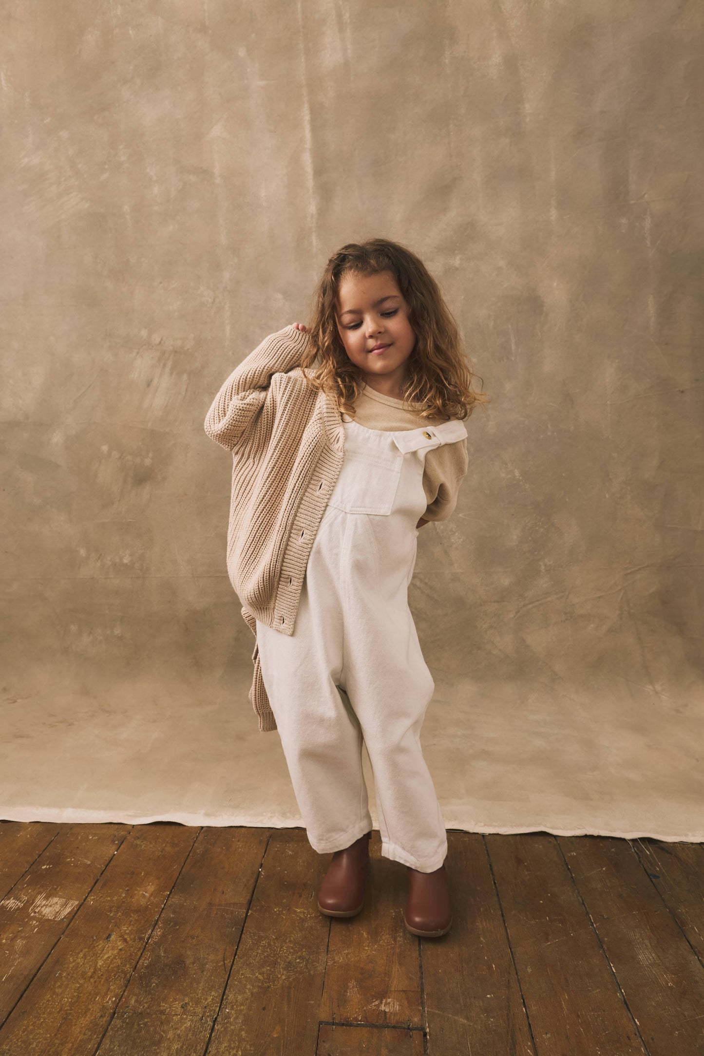 Child wearing a beige cardigan and white overalls standing against a textured brown wall.