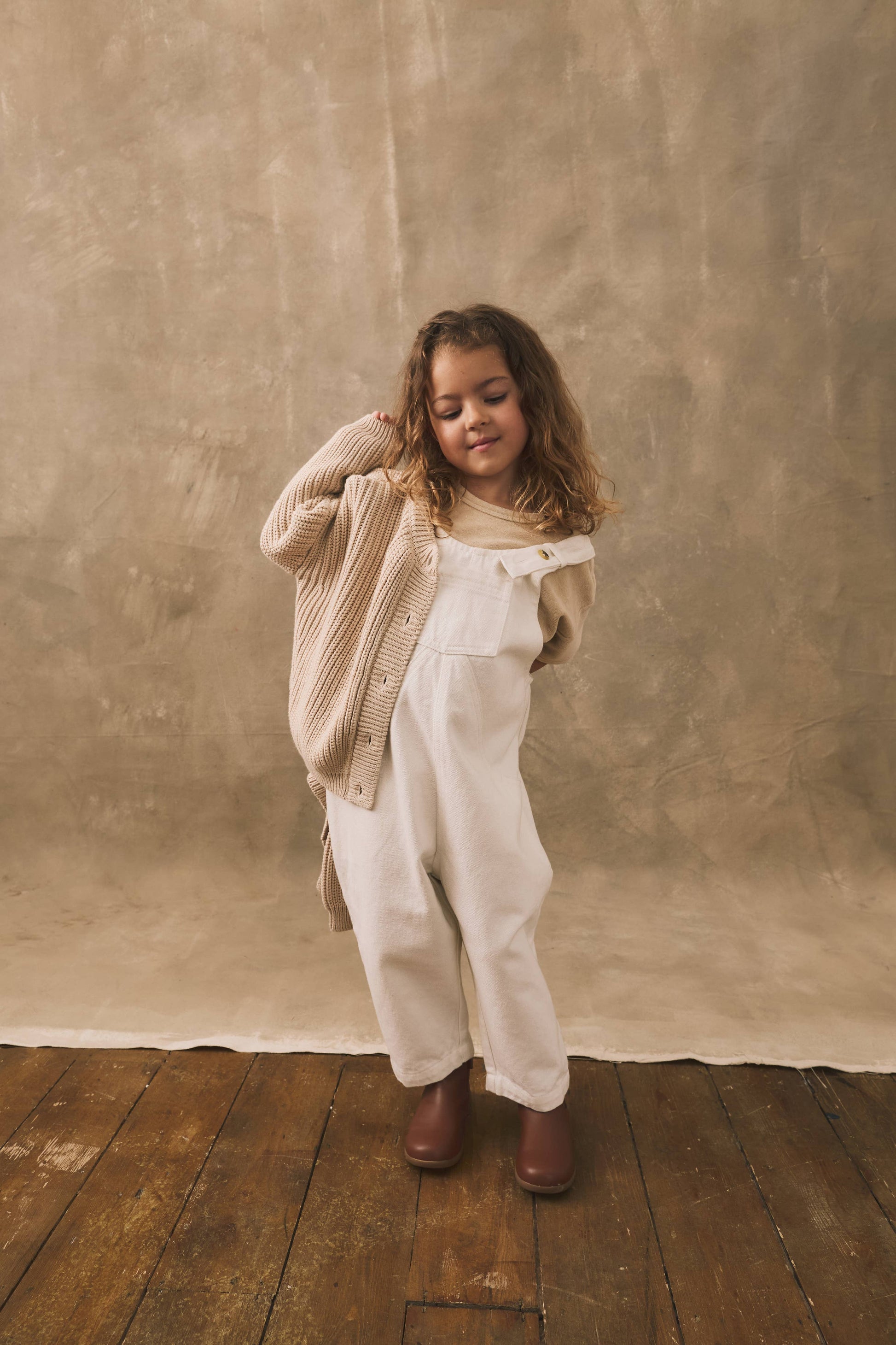 Child wearing a beige cardigan and white overalls standing against a textured brown wall.
