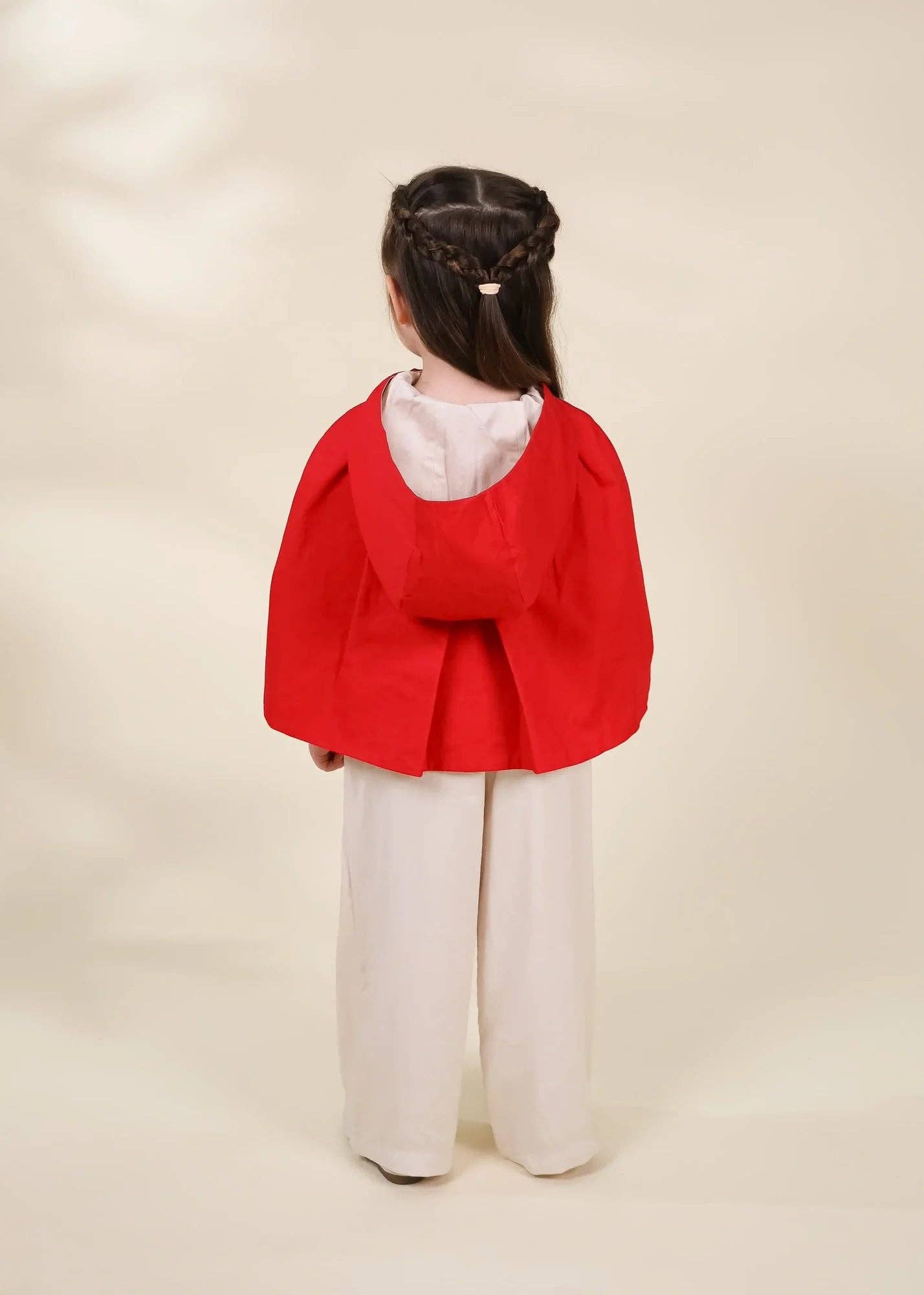 A young girl viewed from behind wearing a bright red hooded cape-style jacket over a white top and white wide-leg pants, standing against a plain warm beige studio background.