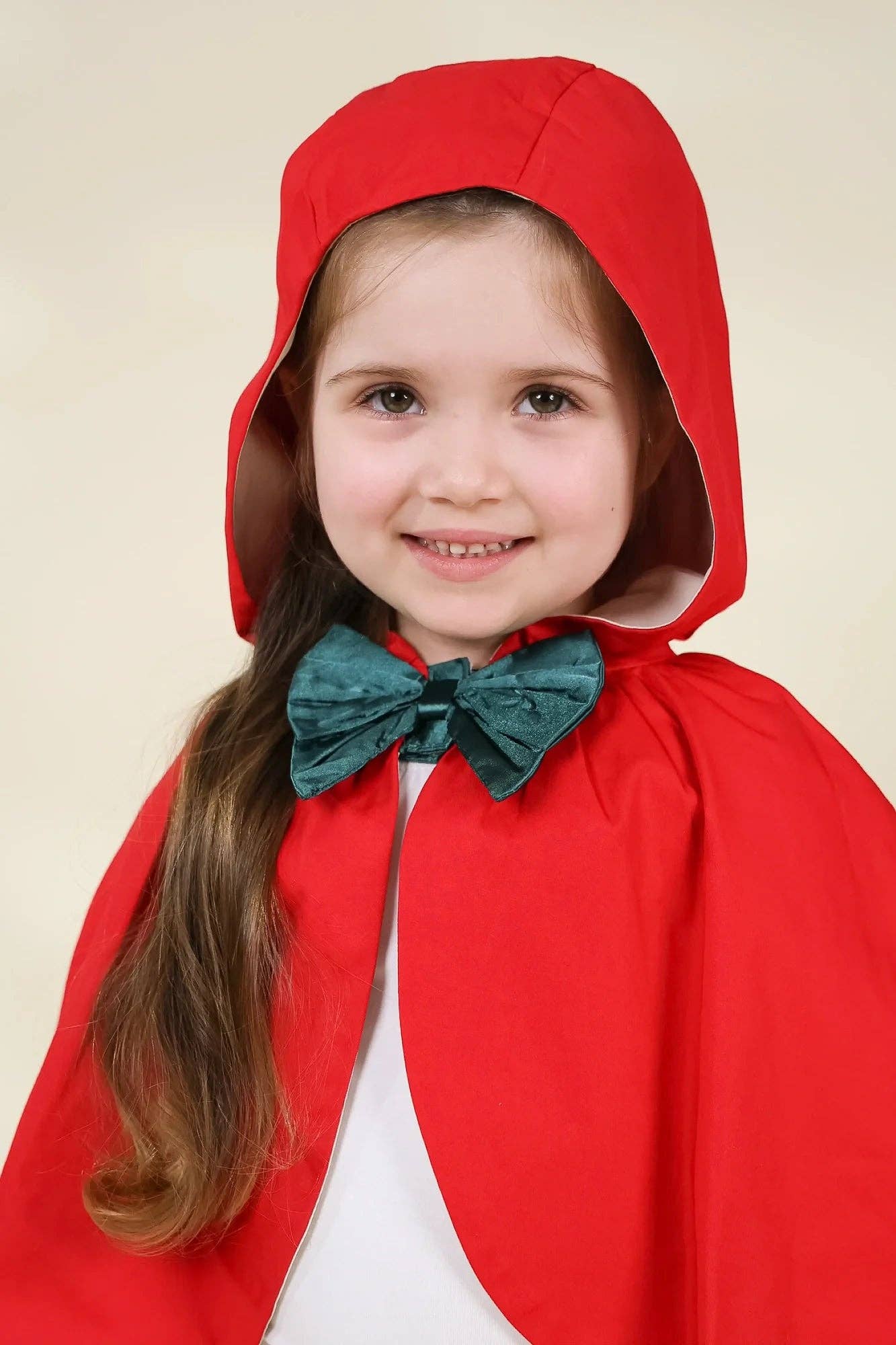 A young girl wearing a red hooded cape and a dark green bow tie over a white shirt, photographed against a plain light beige background.