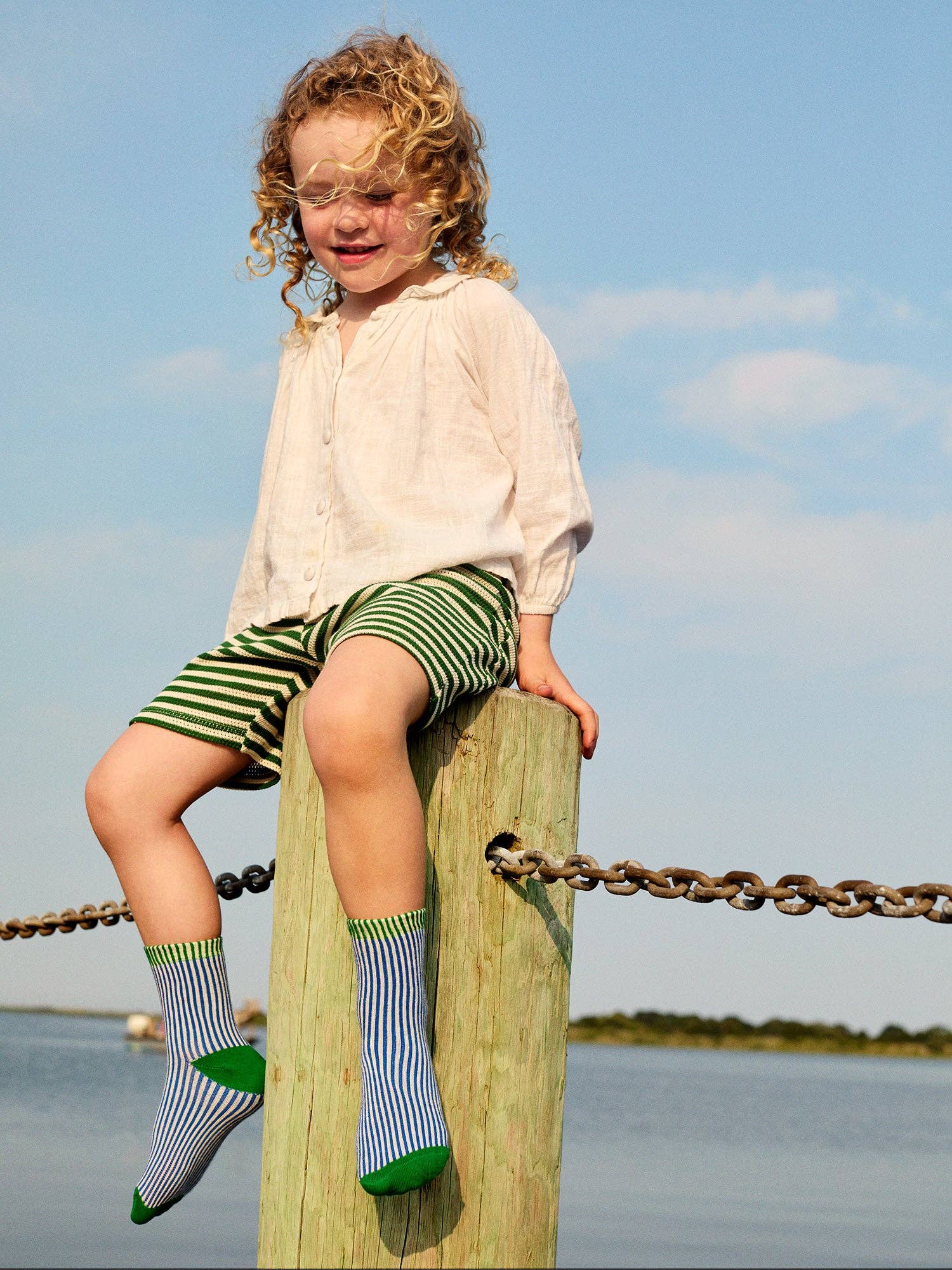 Child sitting on a wooden post by a body of water with a clear blue sky.