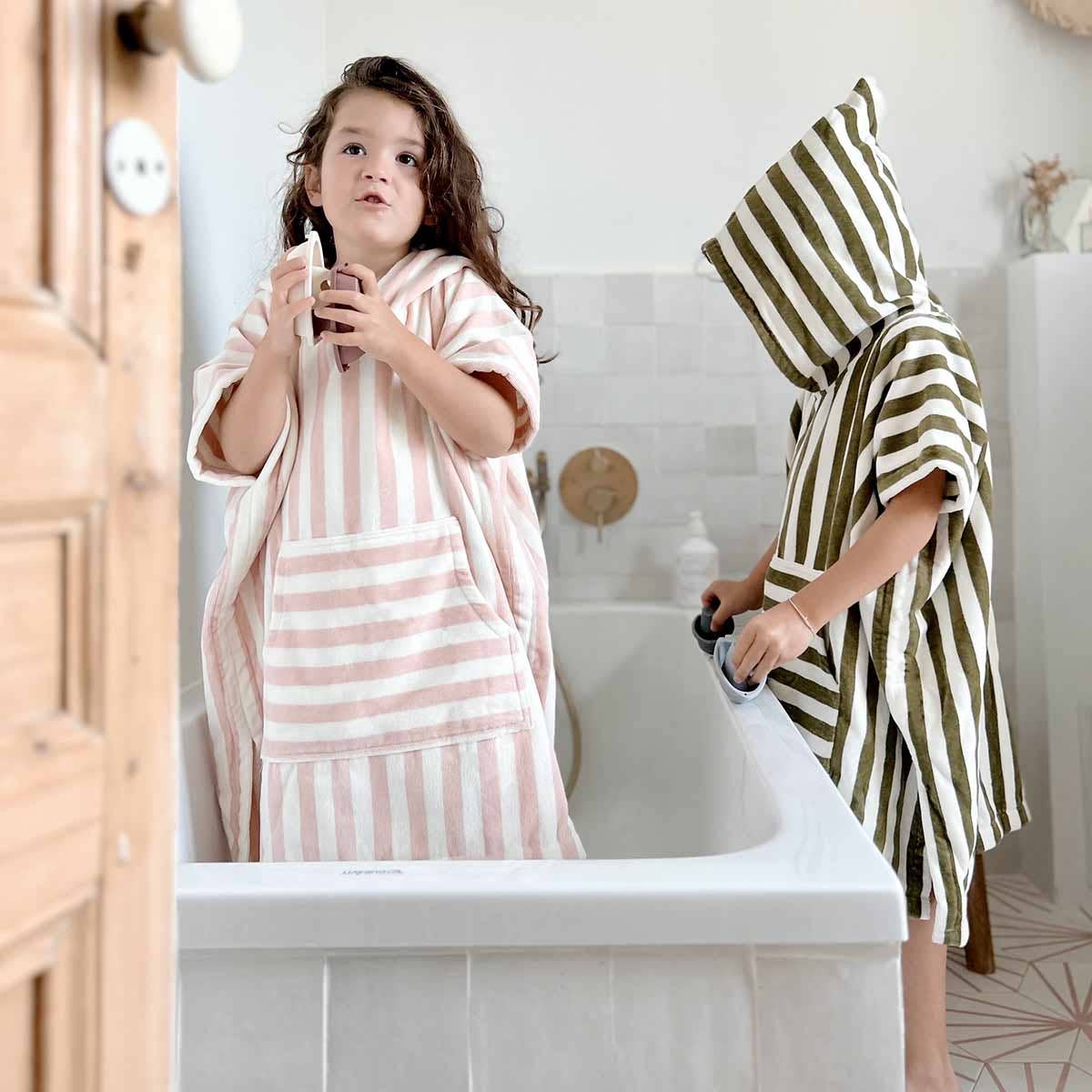 Two children in striped hooded towels standing in a bathroom. Child in pink hooded towel standing in tub with bath toy boats. Other child outside of tub, in green hooded towel.