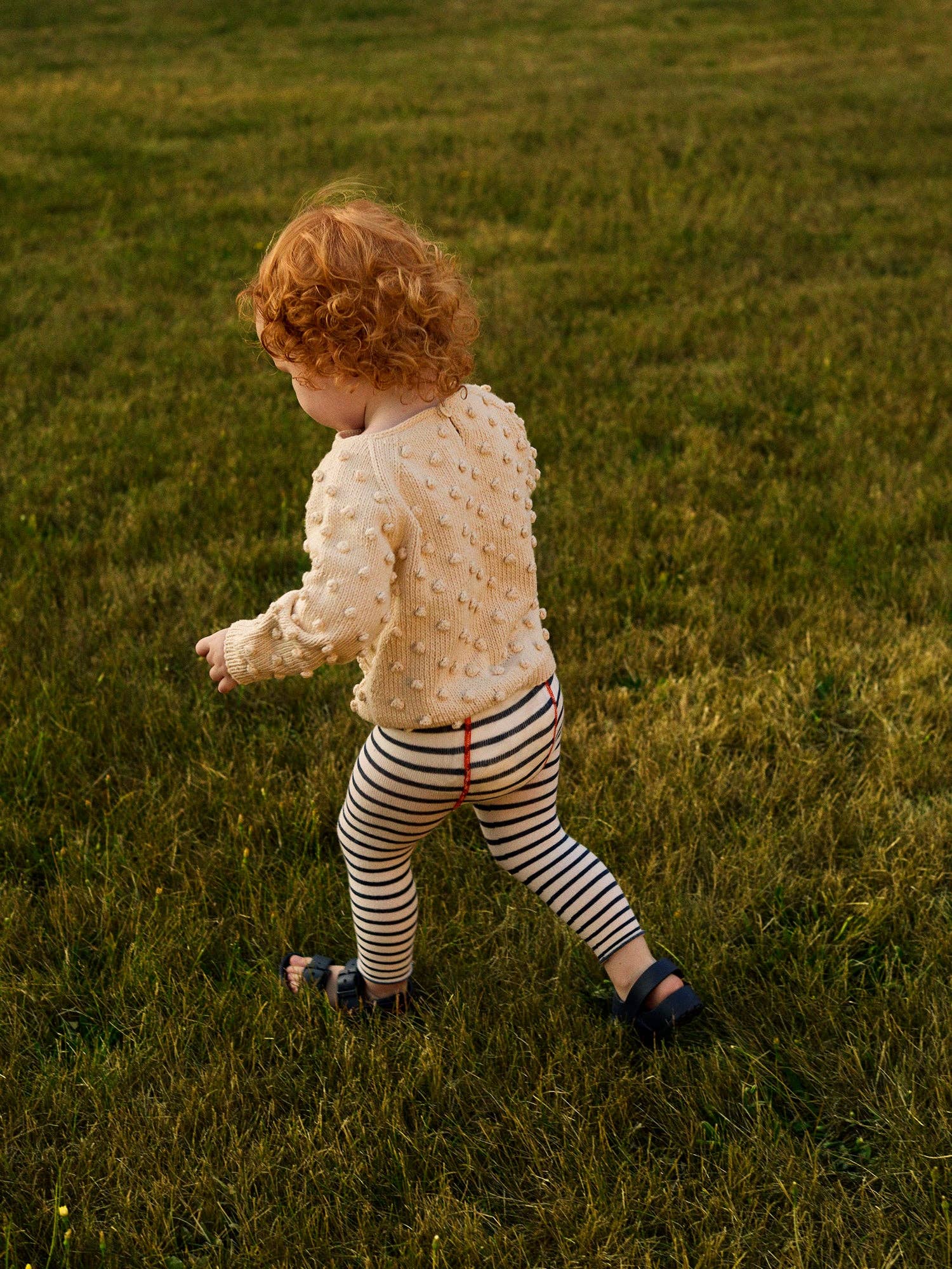 Child walking on grass wearing a textured white sweater and striped pants.