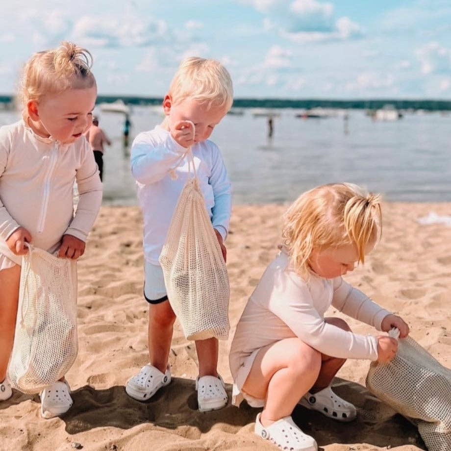 Three children playing on a sunny, sandy beach with mesh bags full of sand toys.