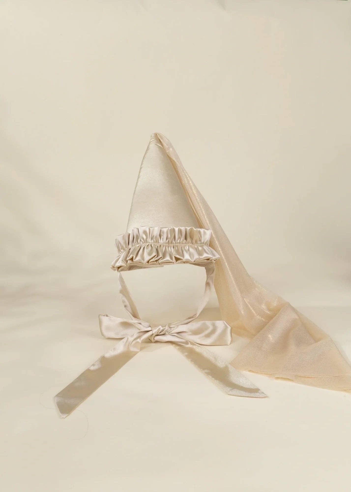 A cream satin fairy hat with a ruffled trim, chin strap, and attached sheer veil, displayed on a neutral cream background.