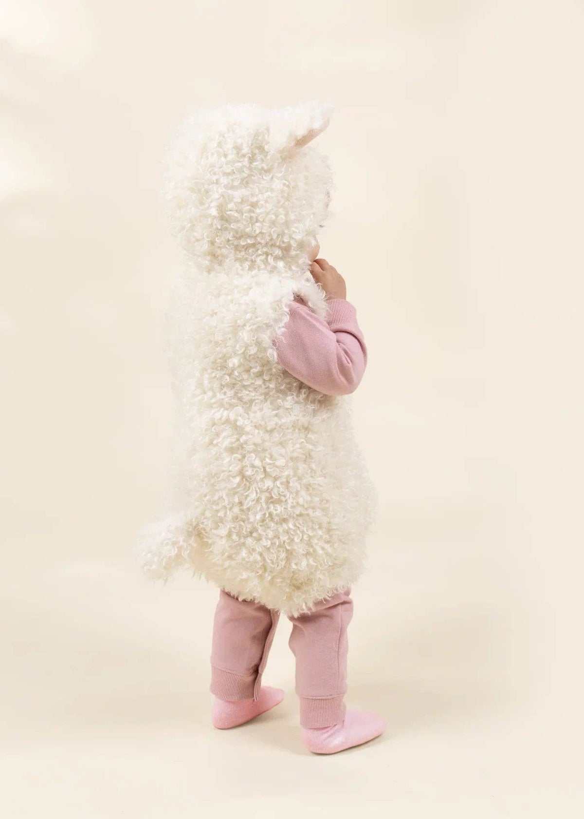 A child wearing a fluffy white sheep costume with a hood, pink long-sleeve top, and pink boot covers, standing against a plain cream background.