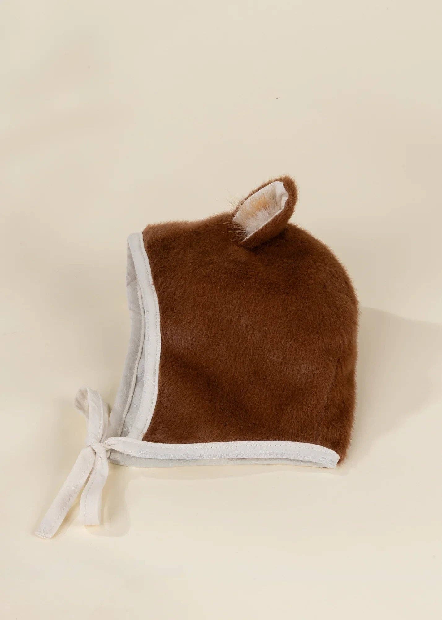 A brown faux fur bonnet hat with white trim and a small white ear detail, tied with a white ribbon on a cream background.