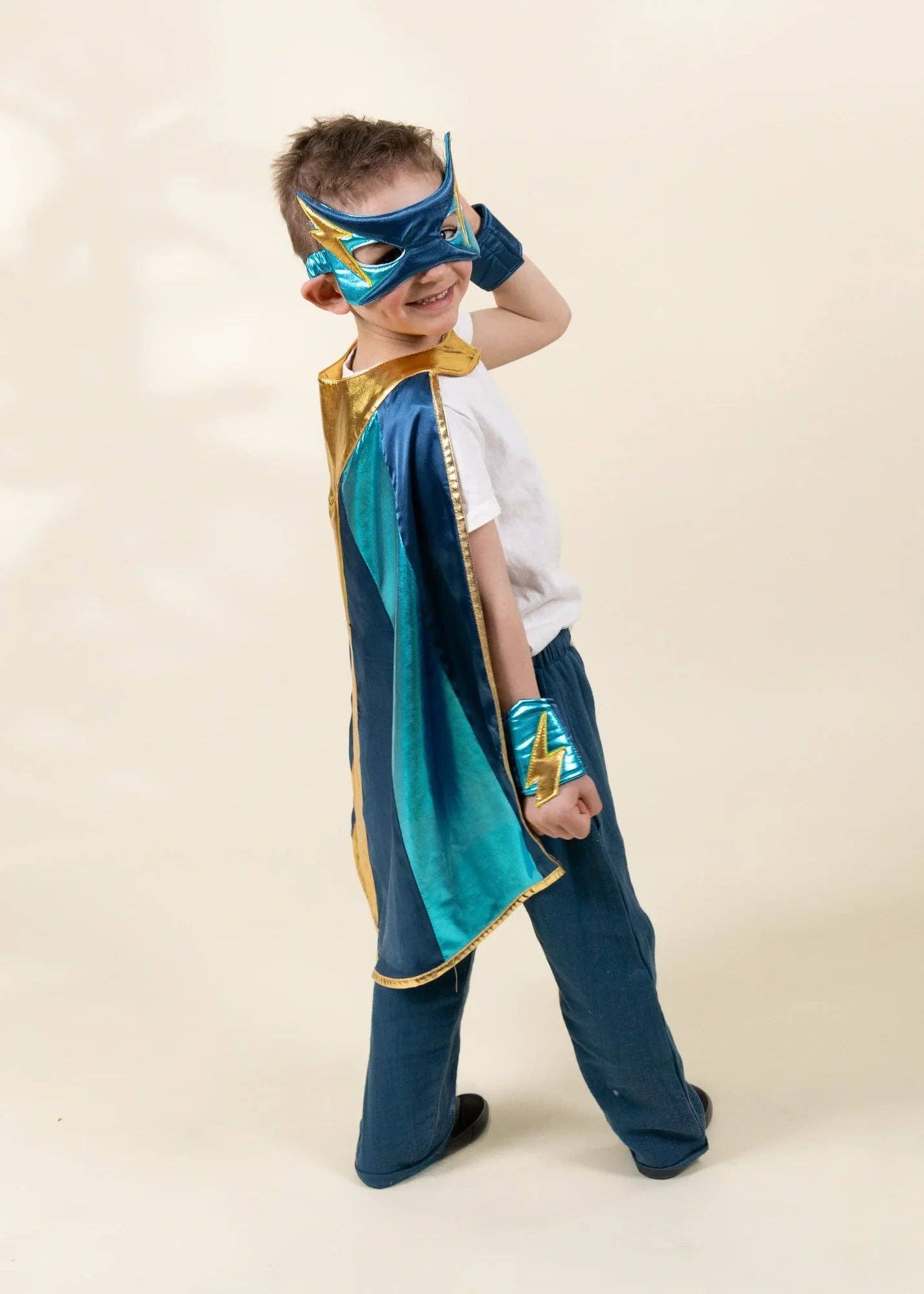 A young boy modeling a superhero costume consisting of a blue and teal cape with gold trim, a matching blue and gold mask, a blue wristband, and dark blue pants, set against a plain beige background.