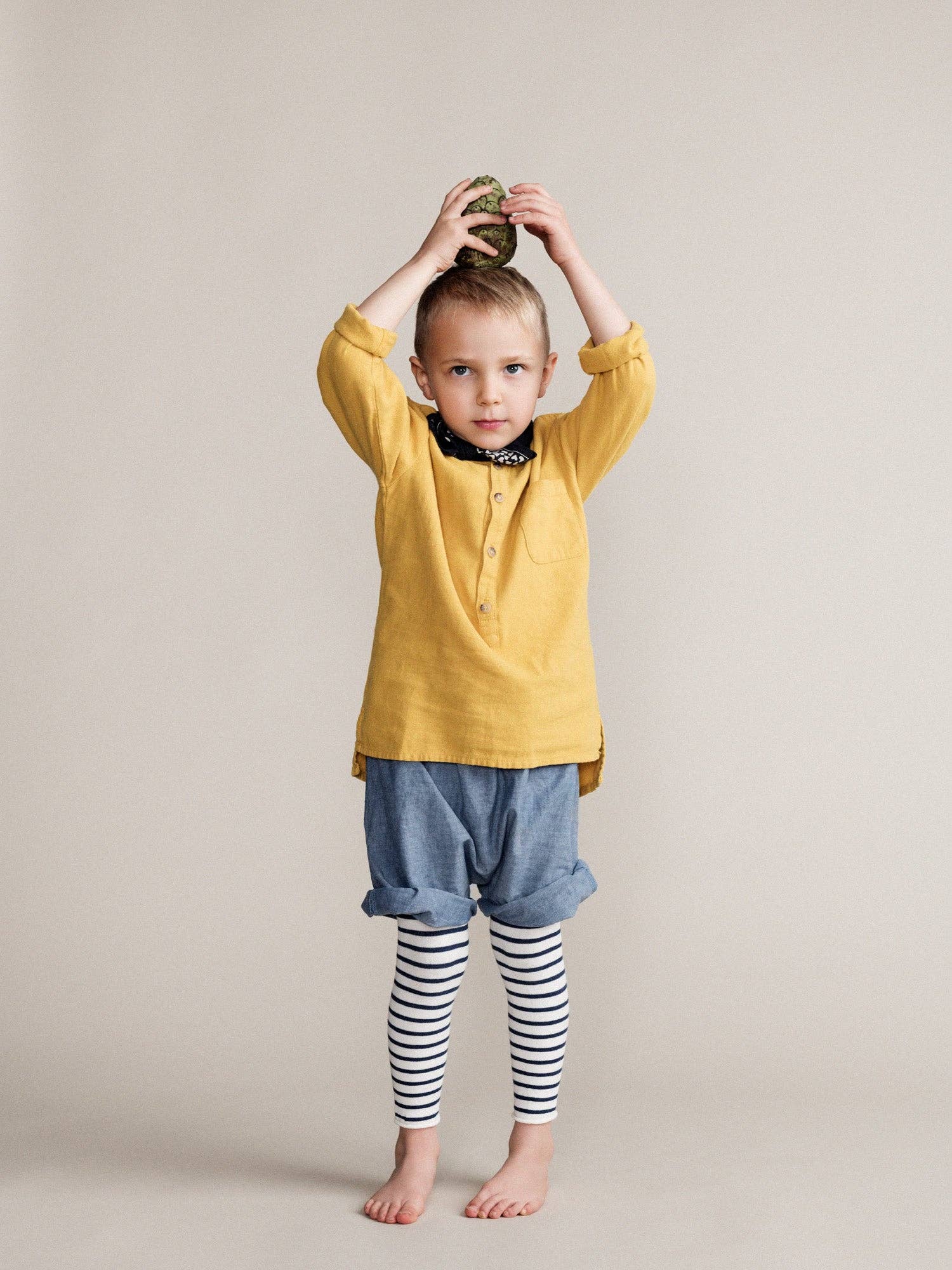 Child wearing a yellow shirt and blue shorts with striped leggings against a plain background