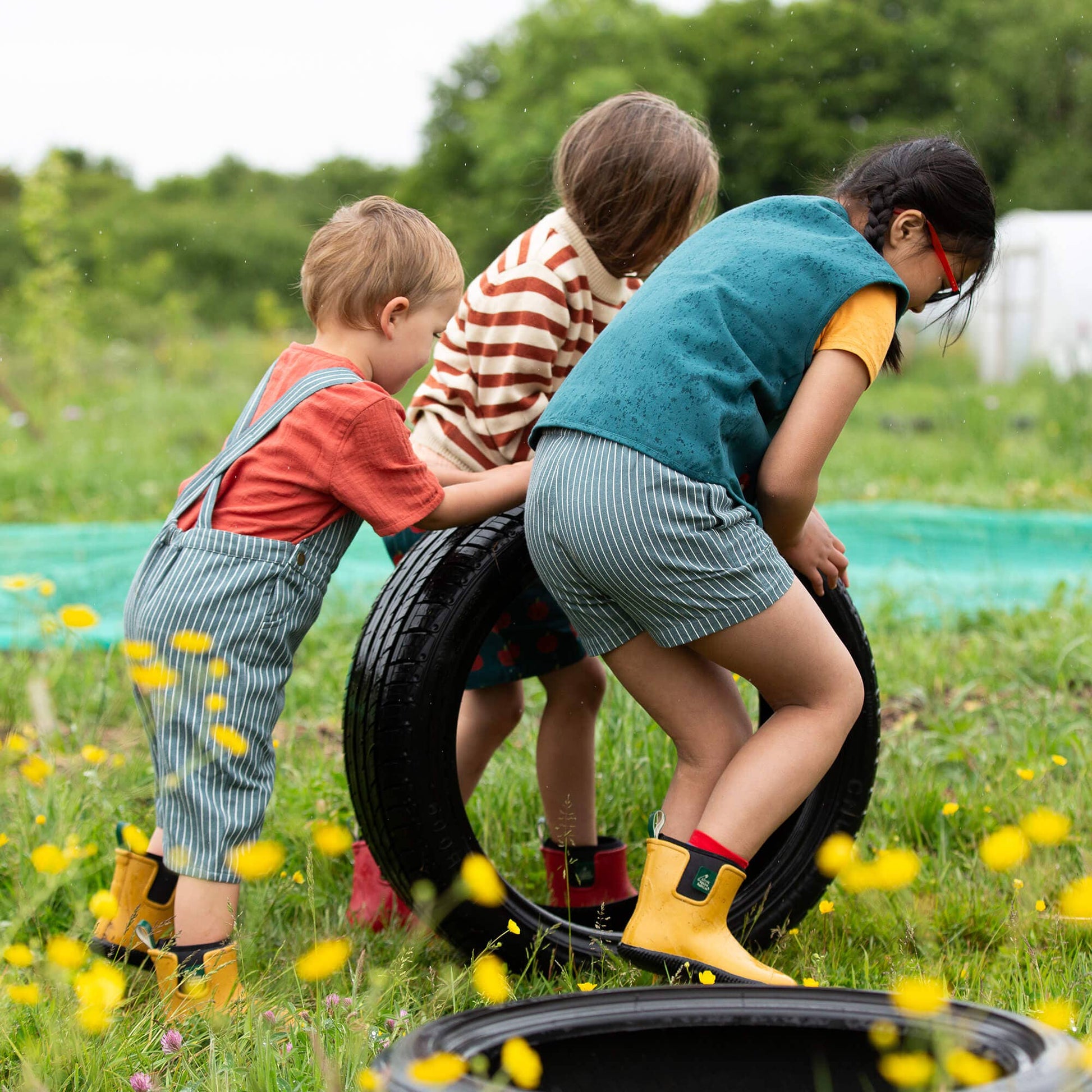 Three children playing with a tire in a grassy field with yellow flowers.