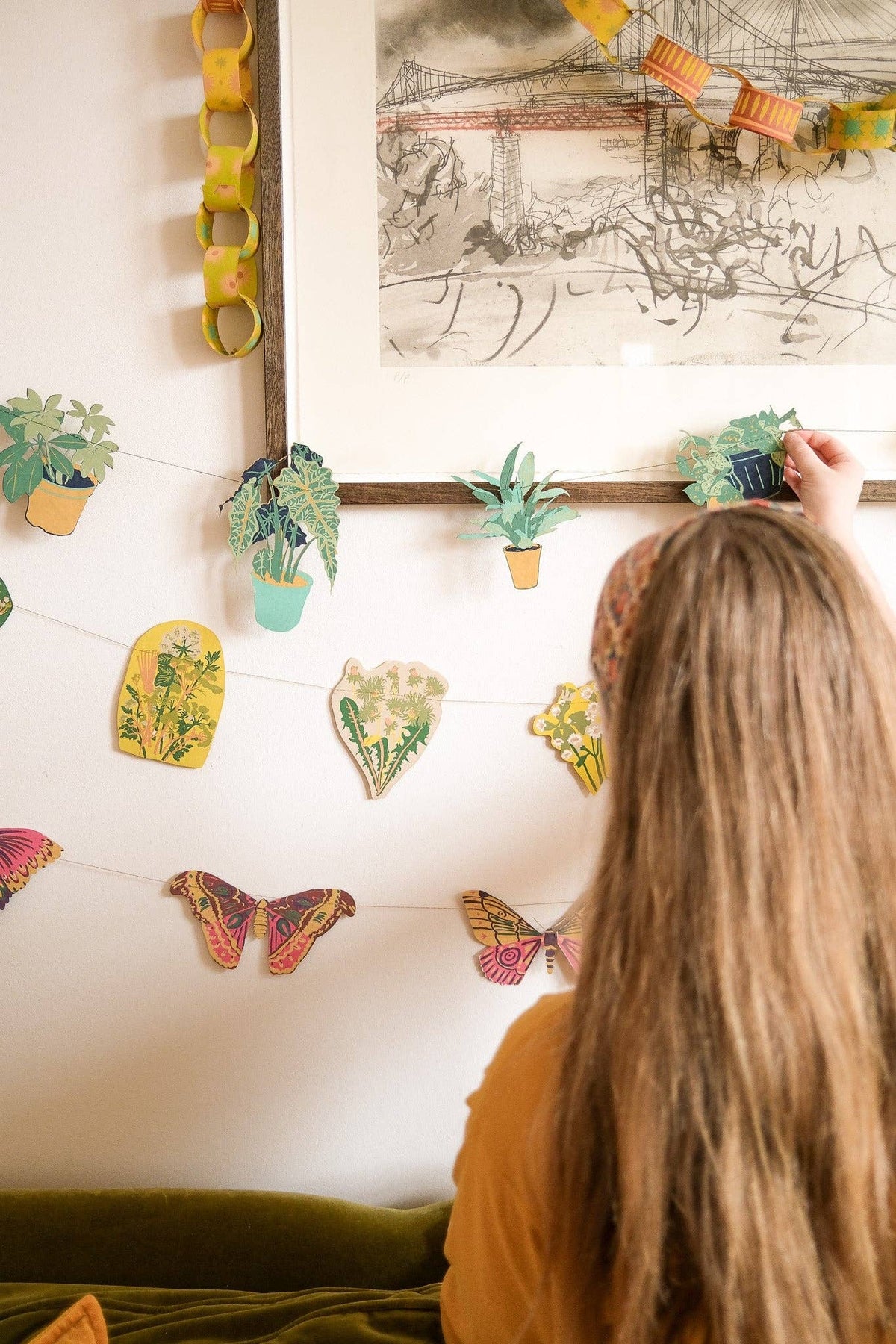 Person arranging paper garlands beneath framed artwork decorated with a yellow paper chain.