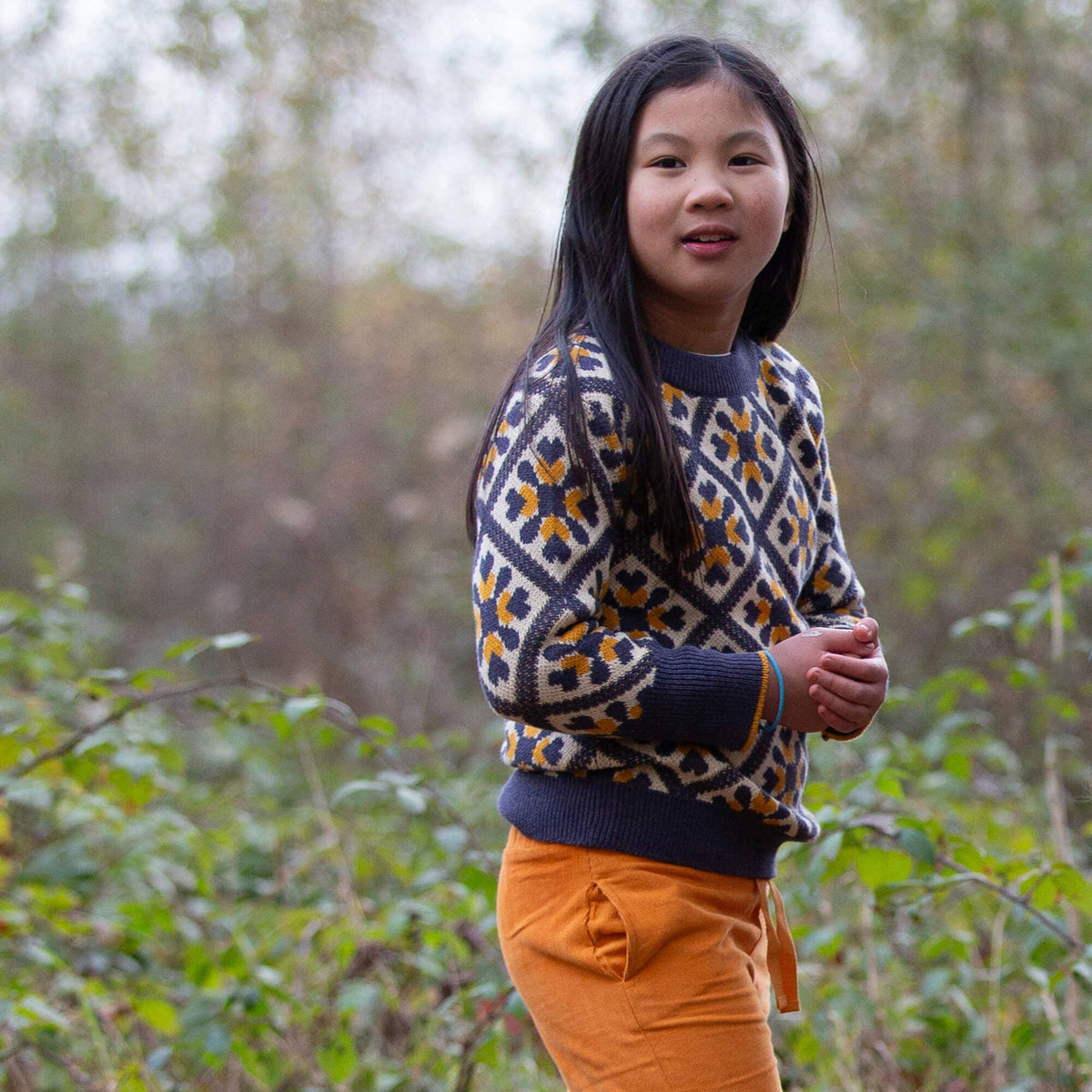 Child wearing a navy, cream and mustard diamond-patterned knitted jumper in woodland.