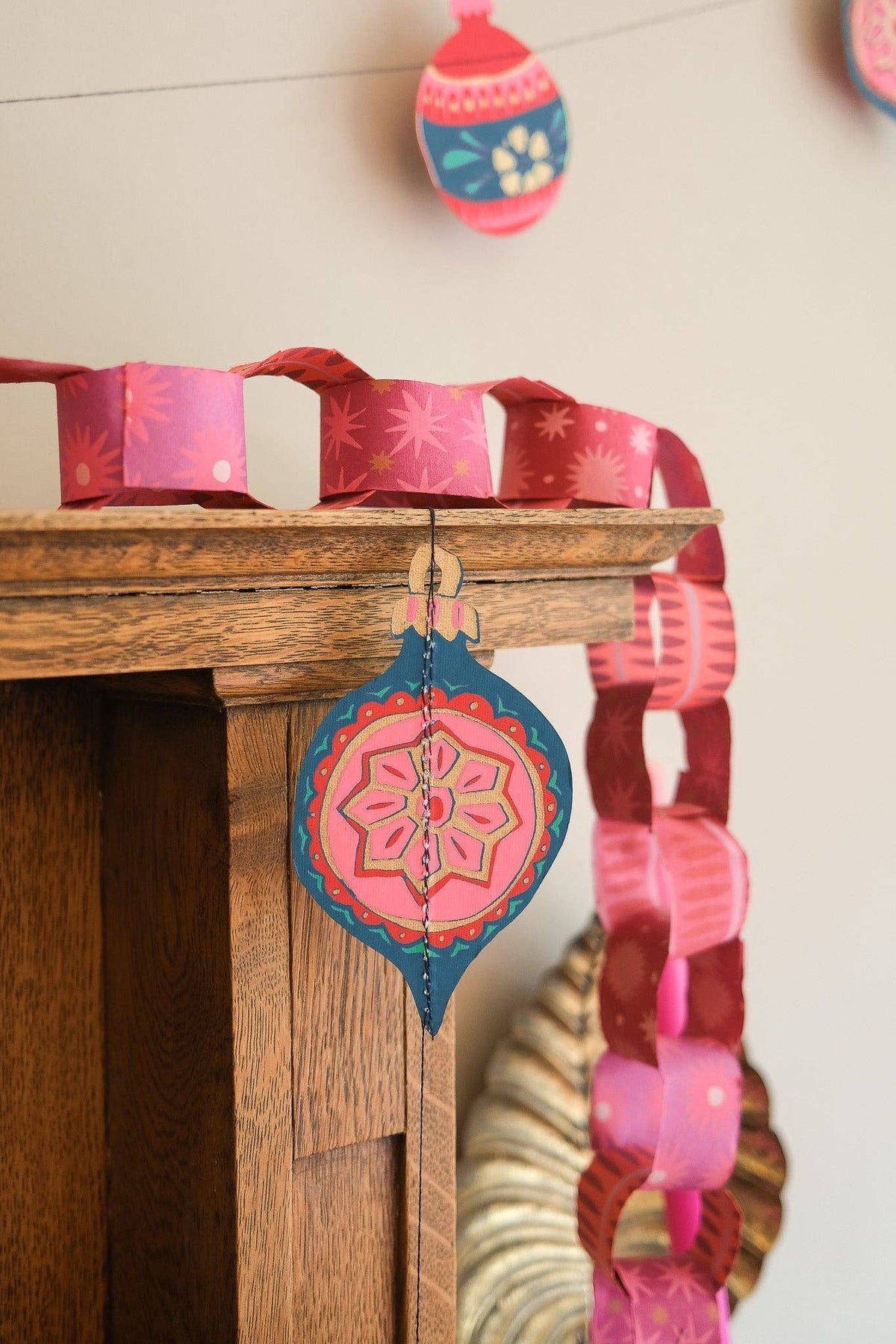 Pink and red paper chain on a wooden mantel beside a colorful paper bauble decoration.
