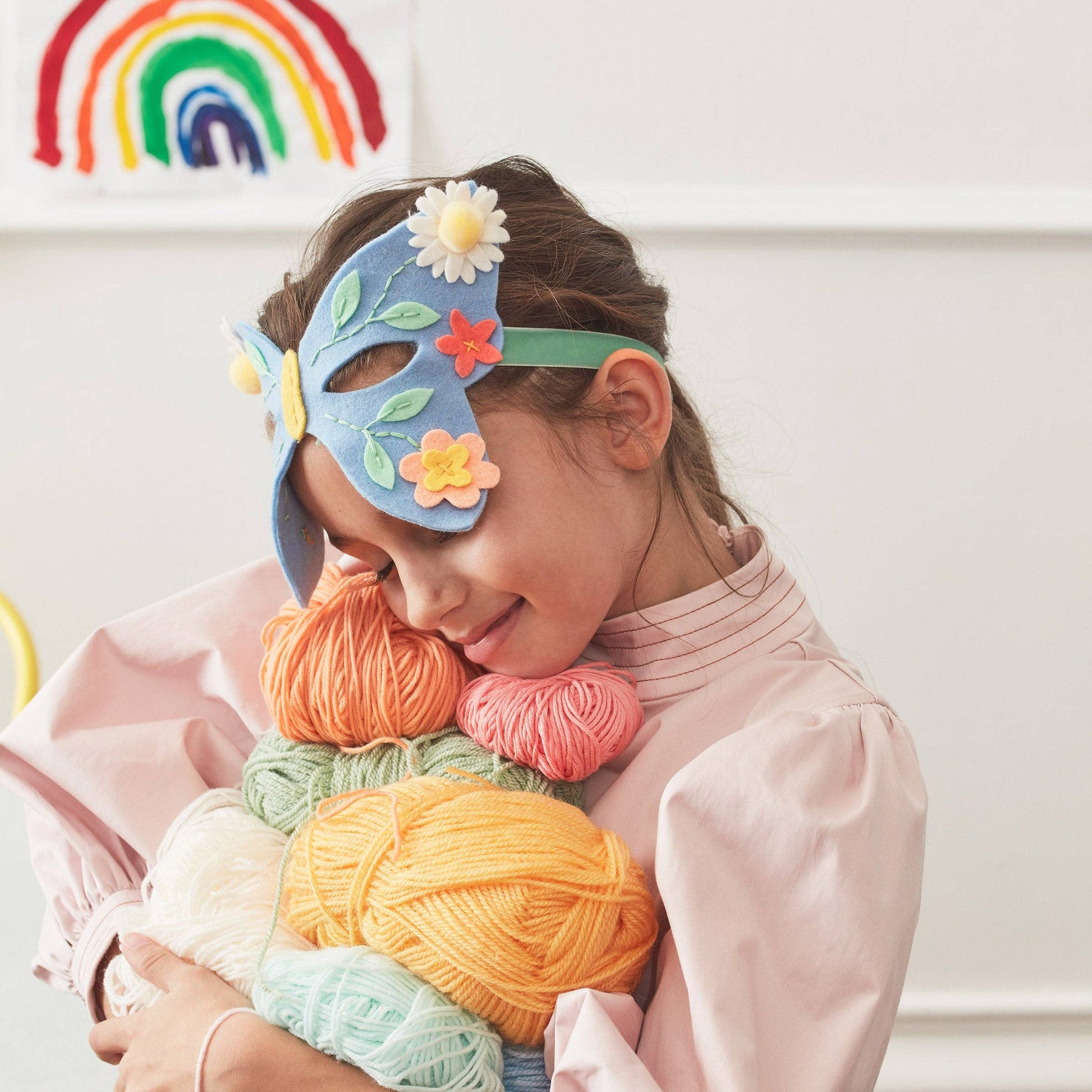 Child wearing a colorful mask holding yarn balls with a rainbow decoration in the background