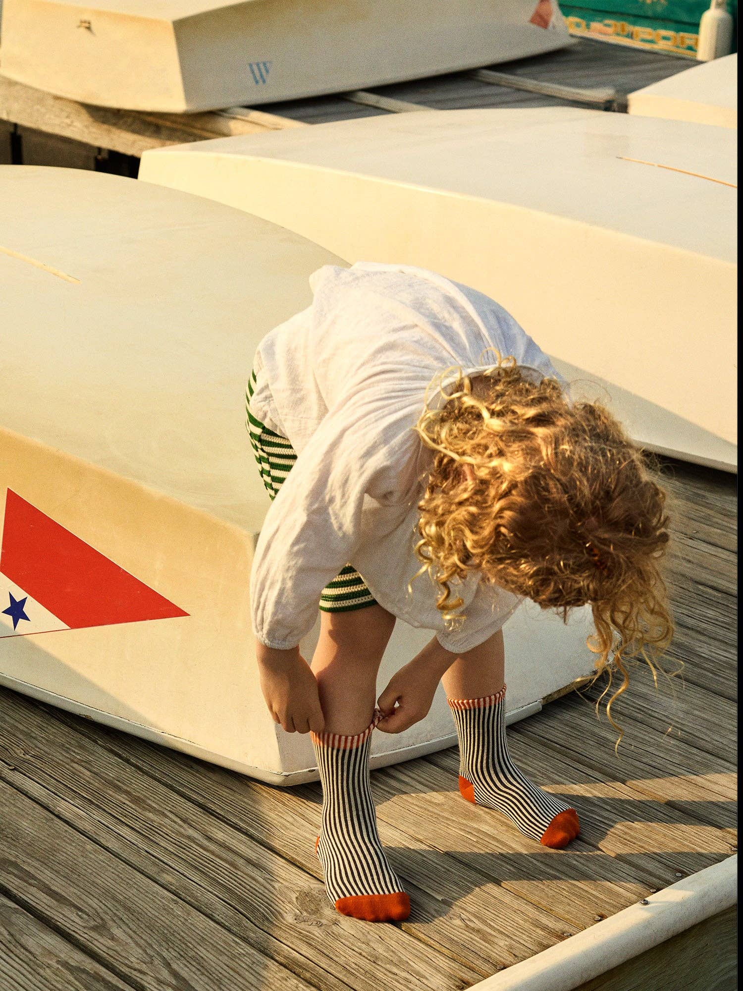Child pulling up black stiped socks while standing on a wooden dock near boats