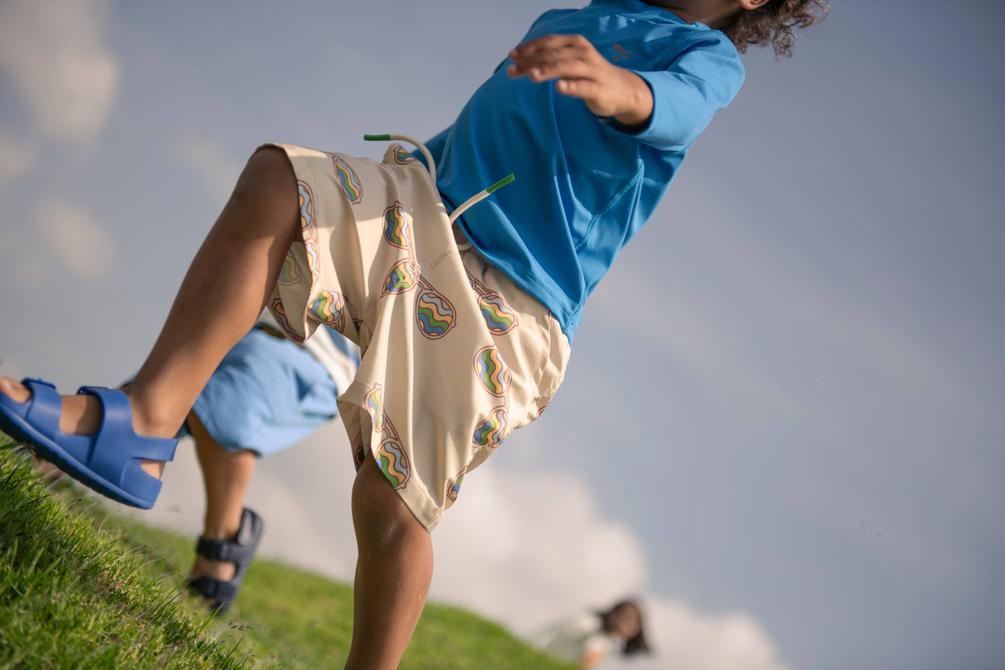 Child in blue rash guard and sandals, with sunglass patterned swim shorts, on grass with a blurred background