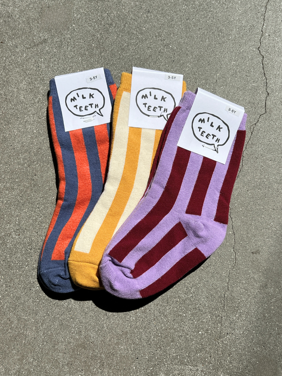 Three pairs of striped socks with 'Milk Teeth' labels on a concrete surface.