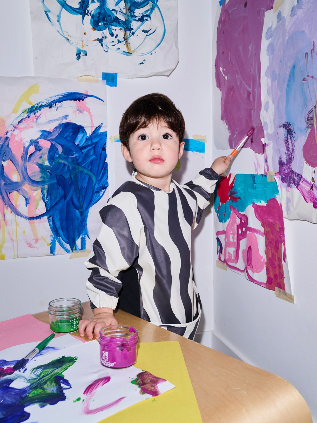 Child in a zebra-striped shirt painting with colorful art on the wall and table.