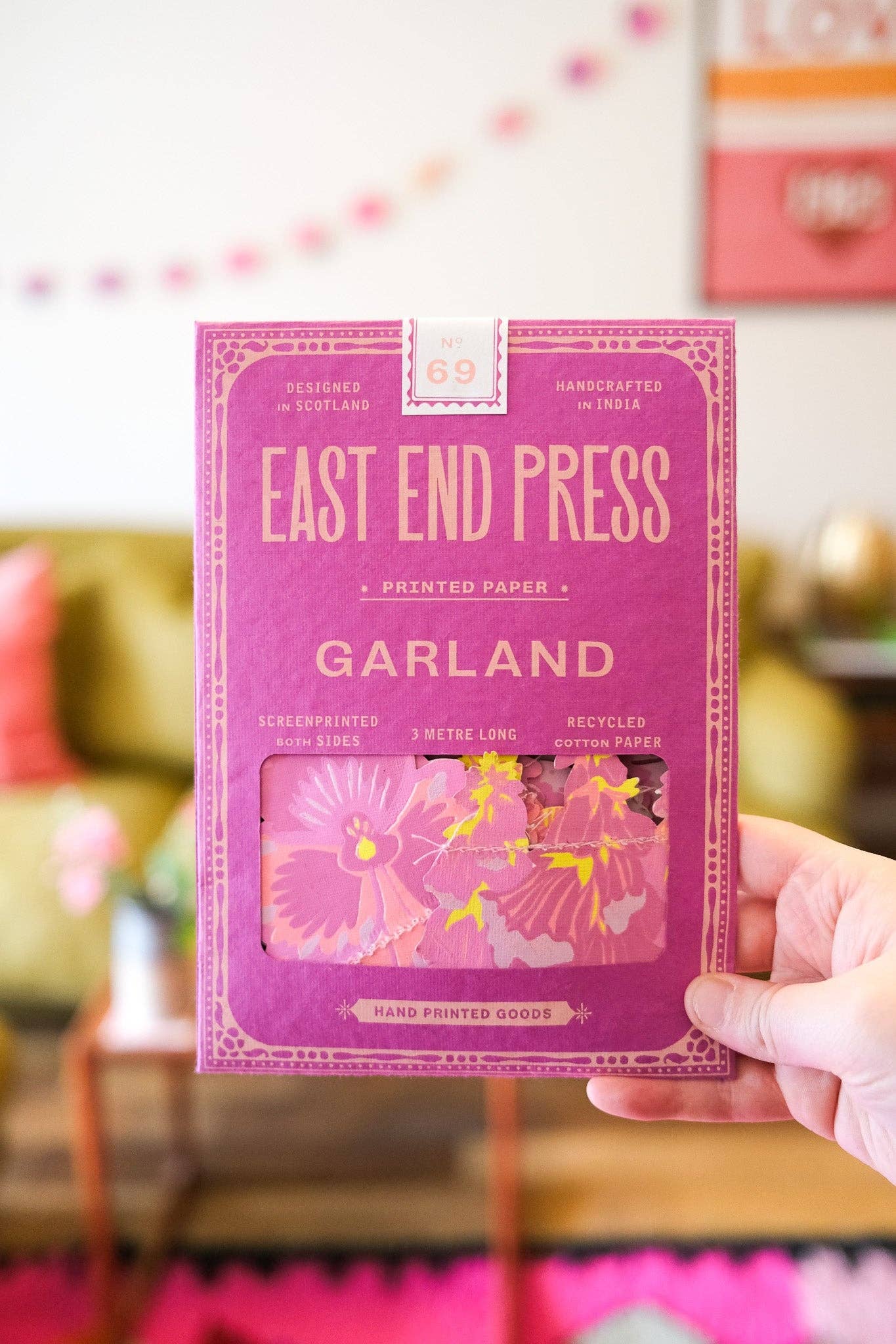 Hand holding a package of East End Press paper Garland with a blurred indoor background.