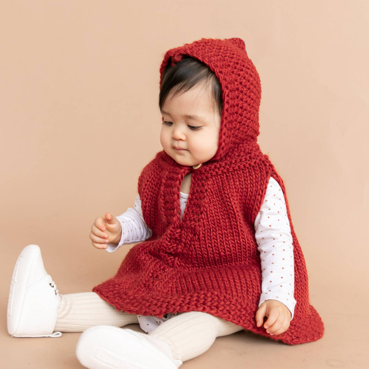 Baby wearing a red knitted hooded vest on a beige background