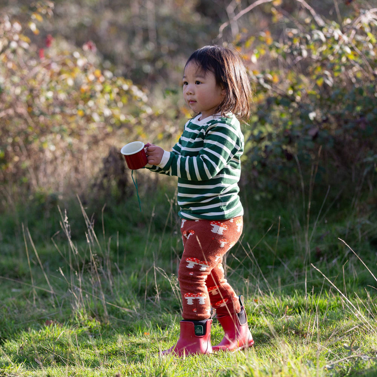 Child wearing rust mushroom joggers and a striped top while holding a red mug outdoors.