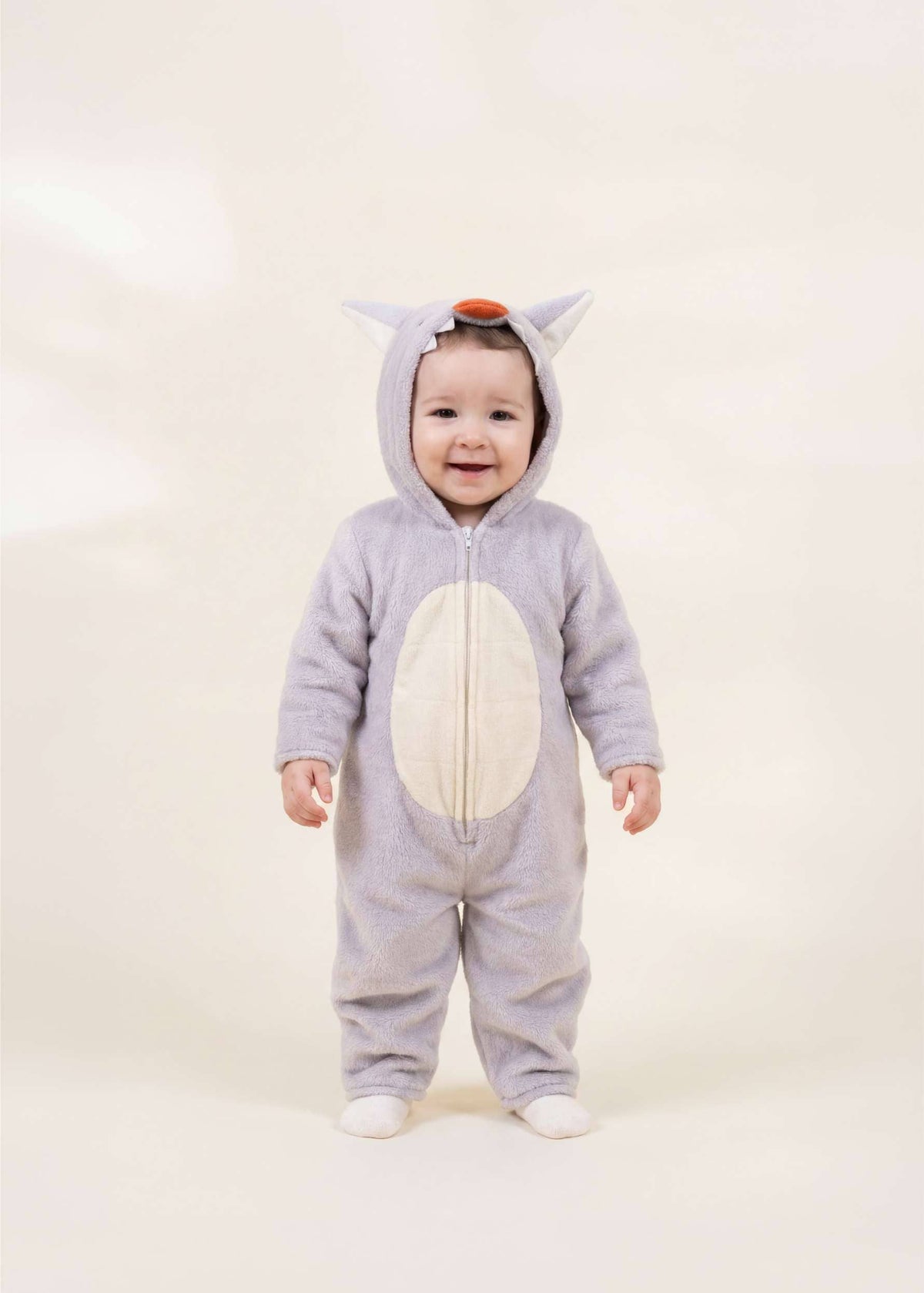 A baby wearing a light gray animal costume with a white belly patch, standing against a plain cream background.