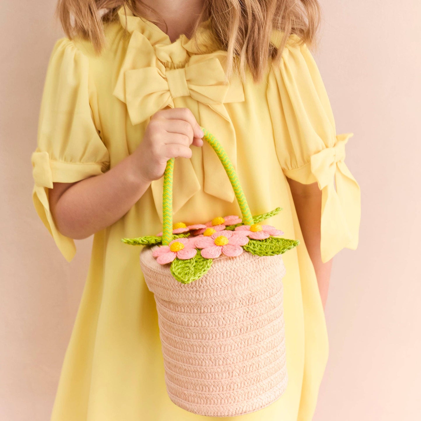 Child in a yellow dress holding a pink woven basket with floral decorations against a light pink background