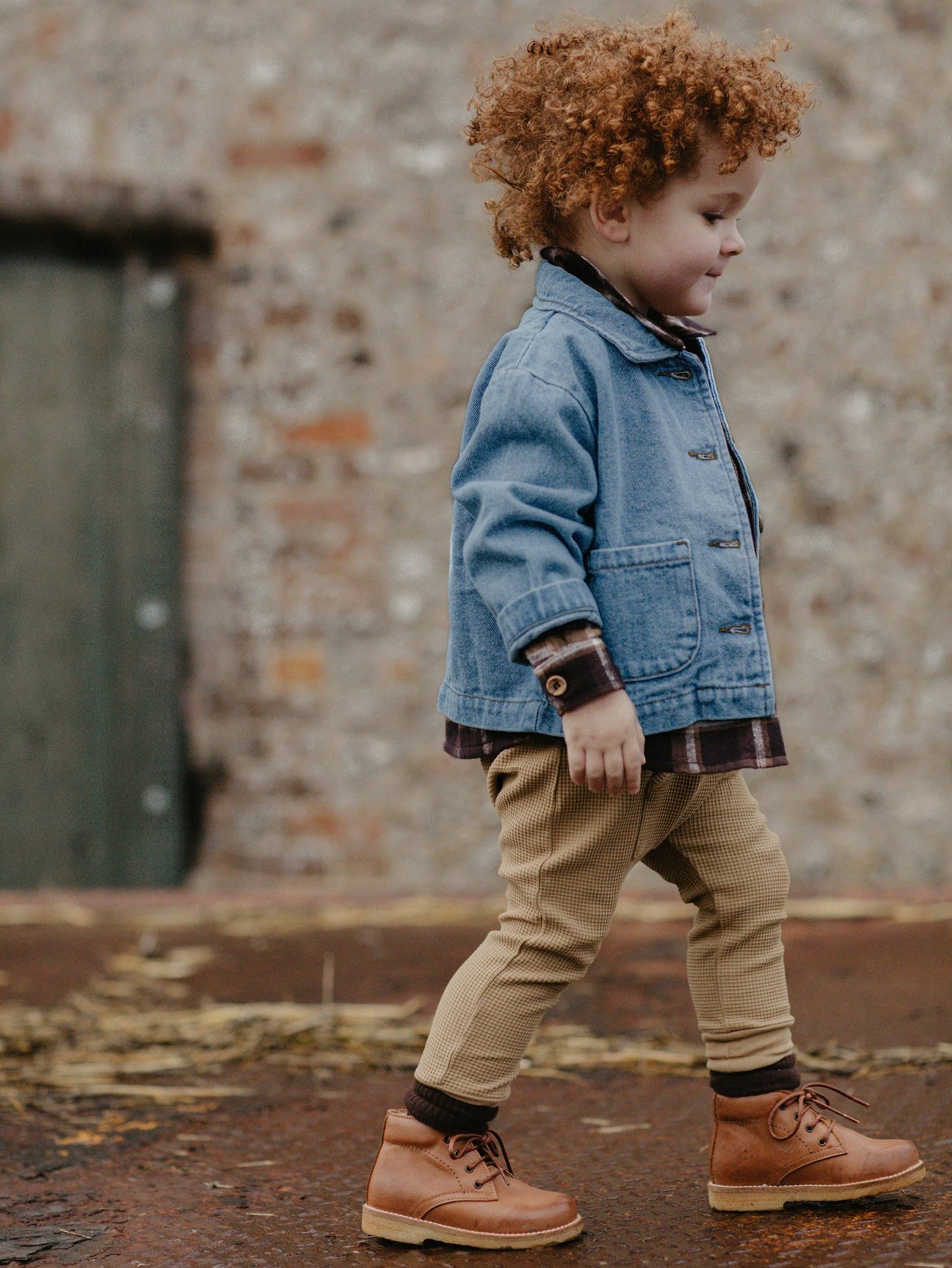 Child wearing a denim jacket and brown boots walking against a brick wall.