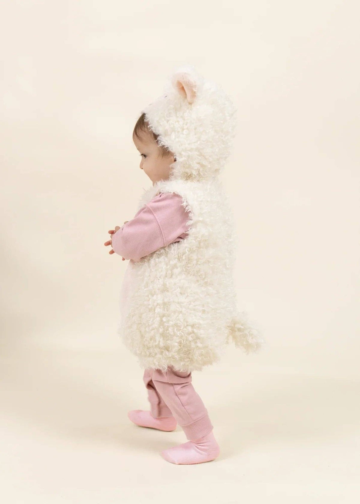 A baby wearing a fluffy white sheep costume with pink sleeves, pink booties, and a pink hood, standing against a solid cream background.