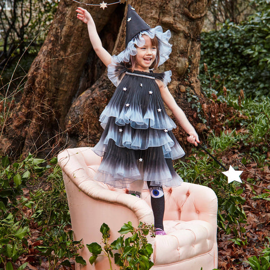 A young girl wearing a black and blue gradient wizard costume stands on a pink armchair in front of a large tree outdoors, holding a star wand.