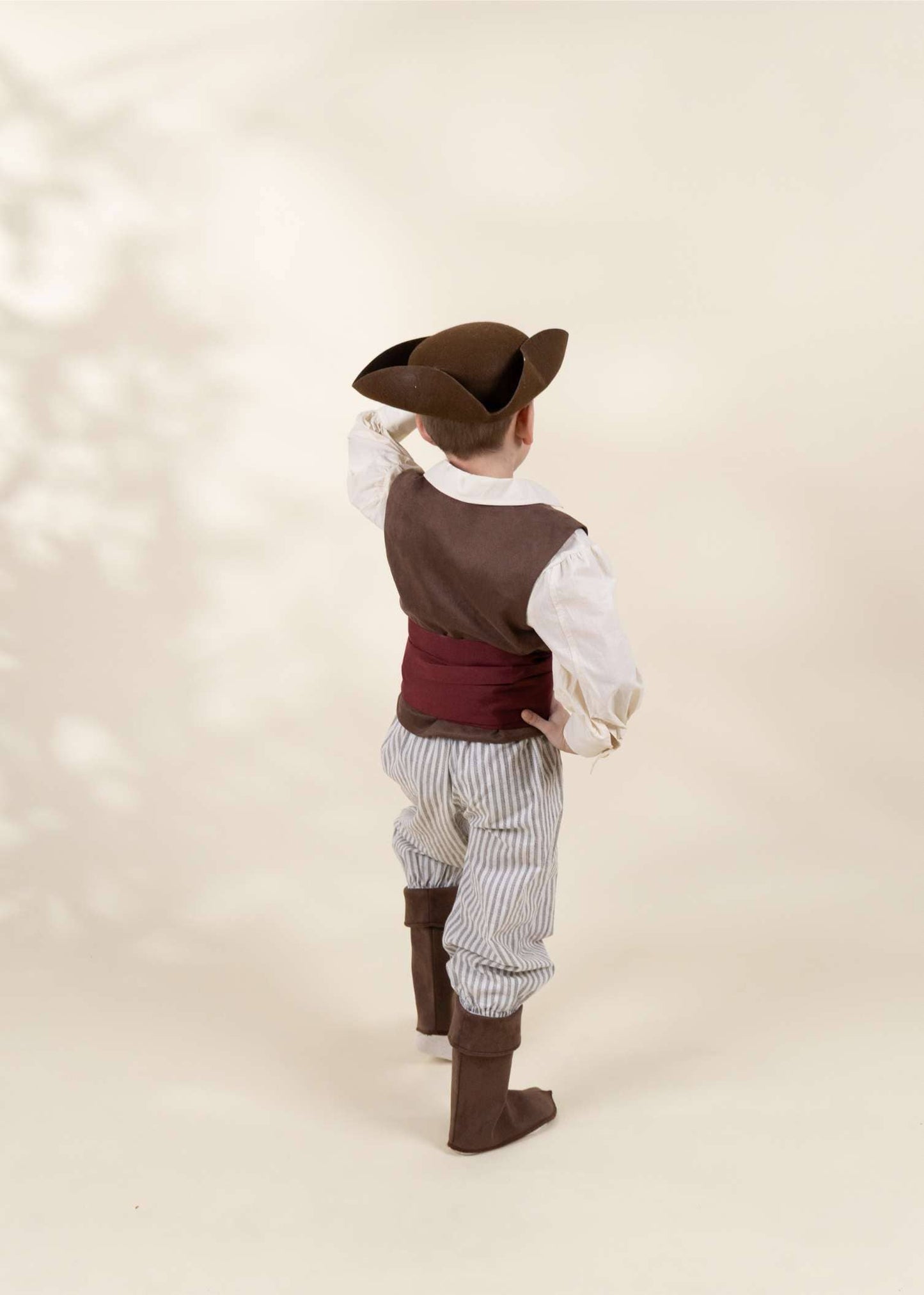 A child wearing a pirate costume, photographed from behind against a warm beige studio background with soft leaf shadows. The costume includes a brown hat, brown vest, white shirt, dark red waist cloth, striped pants, and brown boots.