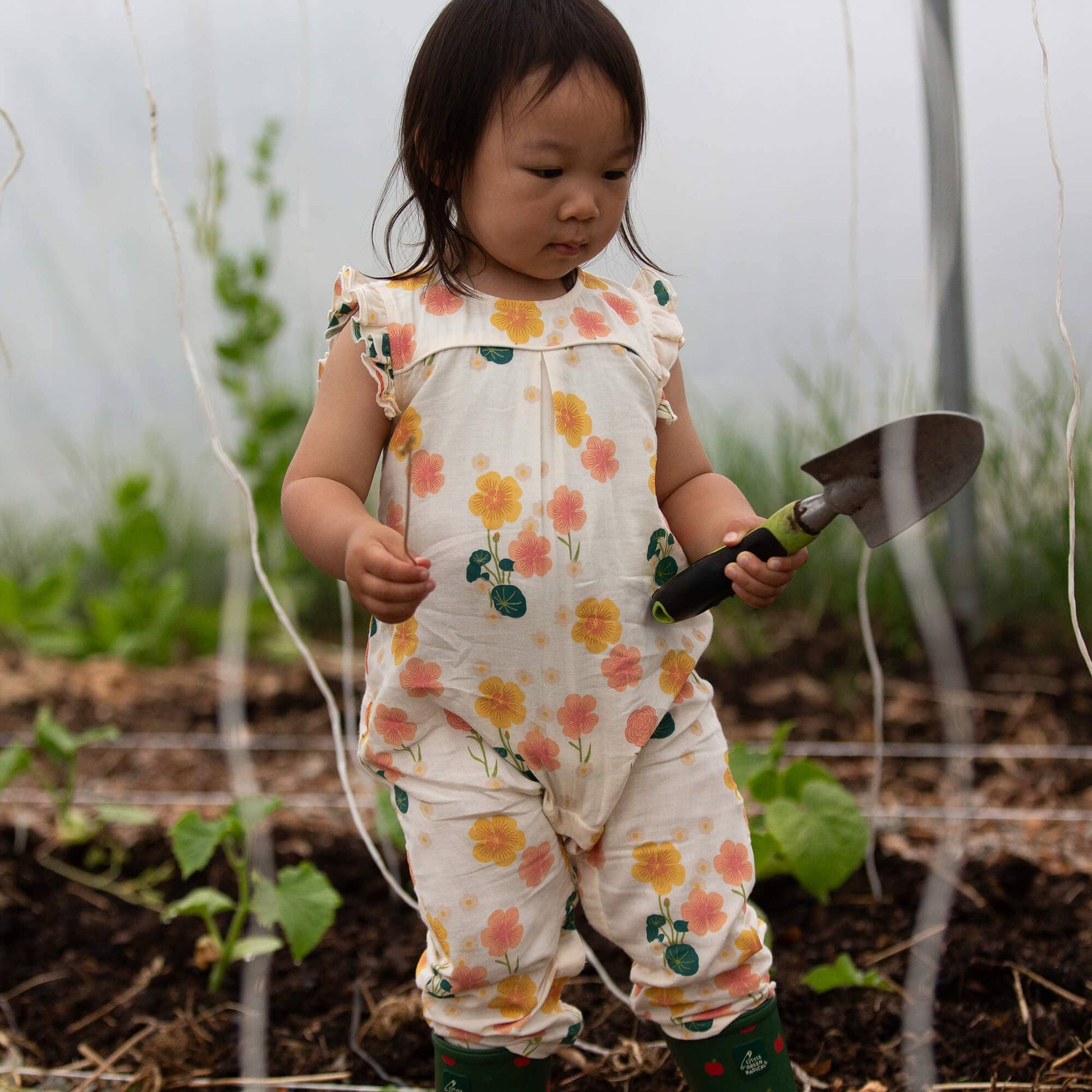 Child in a floral romper holding a small gardening tool in a garden setting
