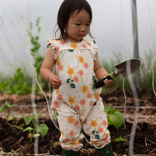 Child in a floral romper holding a small gardening tool in a garden setting