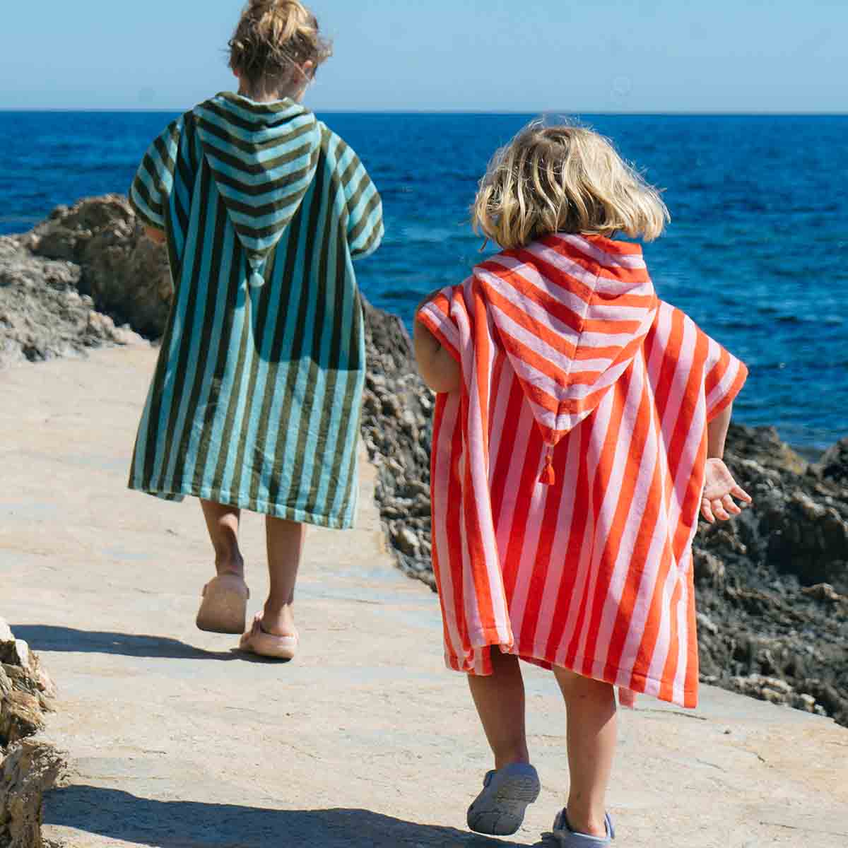 Two children wearing striped hooded towels walking along a rocky path by the ocean.