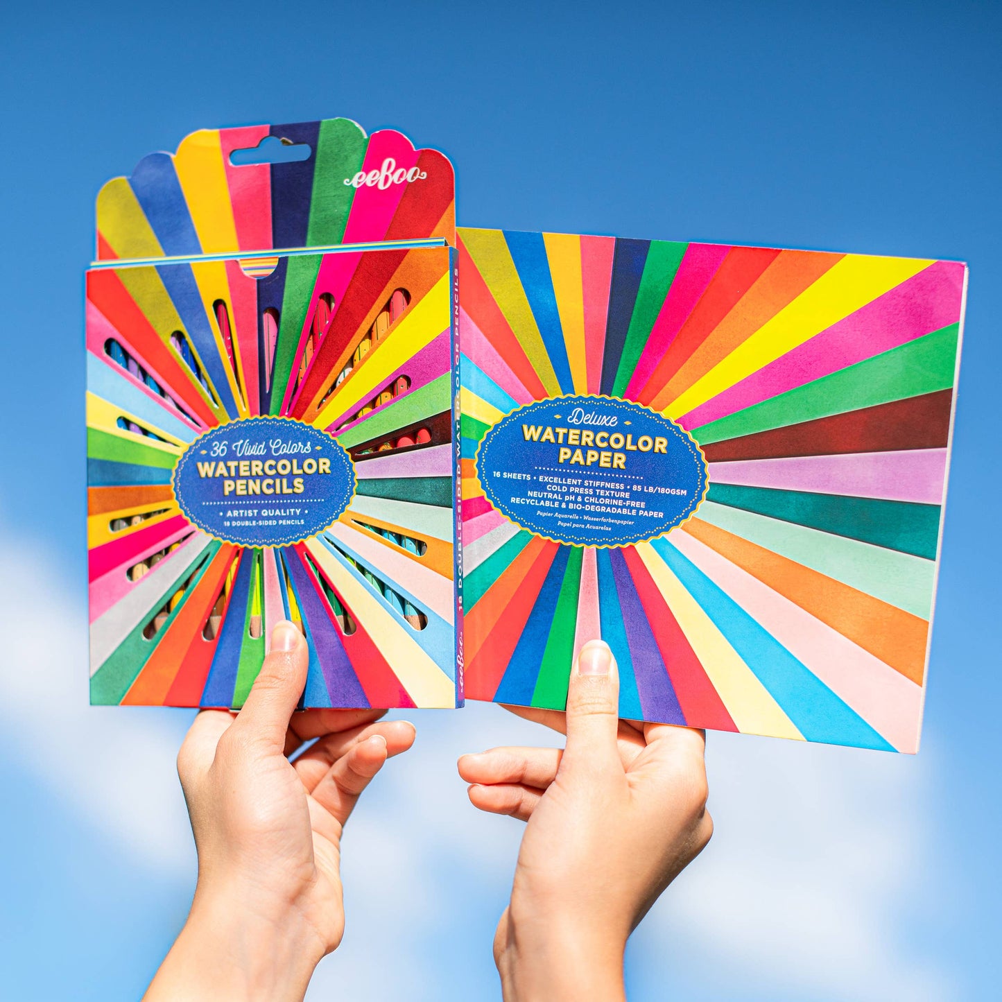 Two packages of watercolor pencils and paper held against a blue sky background
