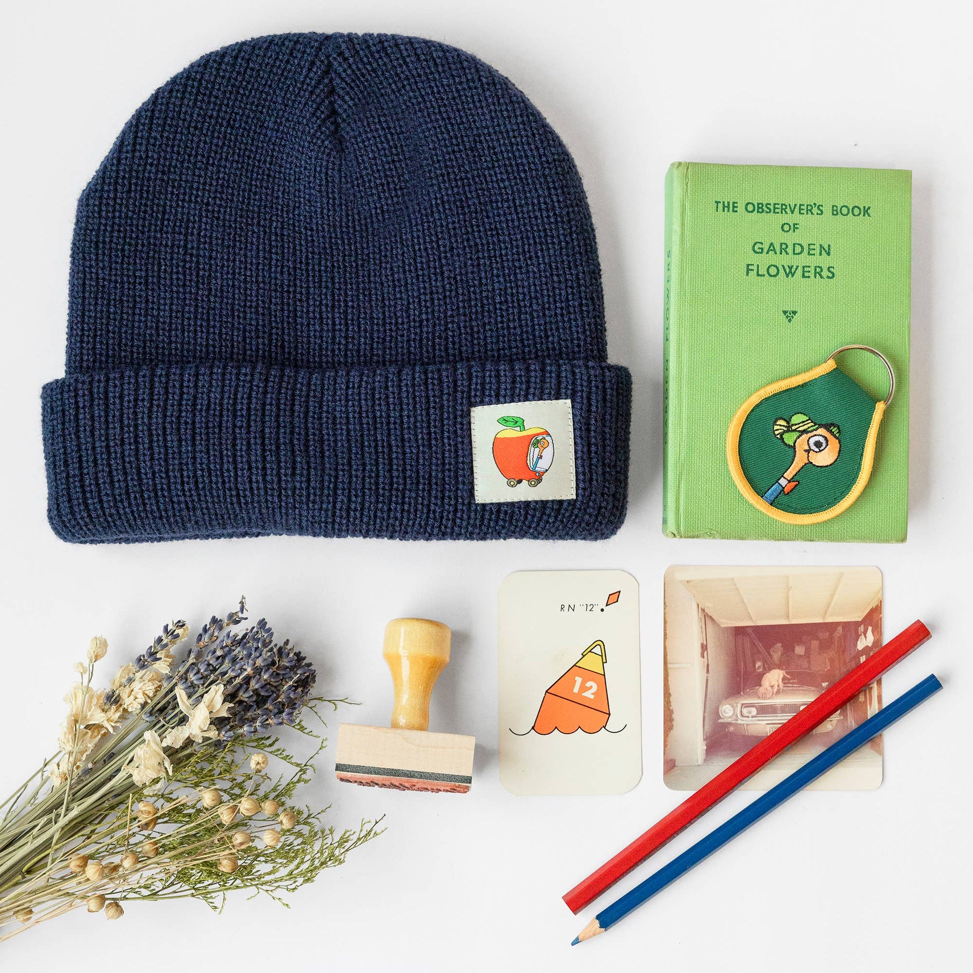 Navy beanie with a patch, green book, stamps, cards, and pencils on a white background