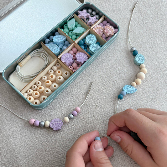 Beaded necklace being crafted with a container of assorted beads on a textured surface.