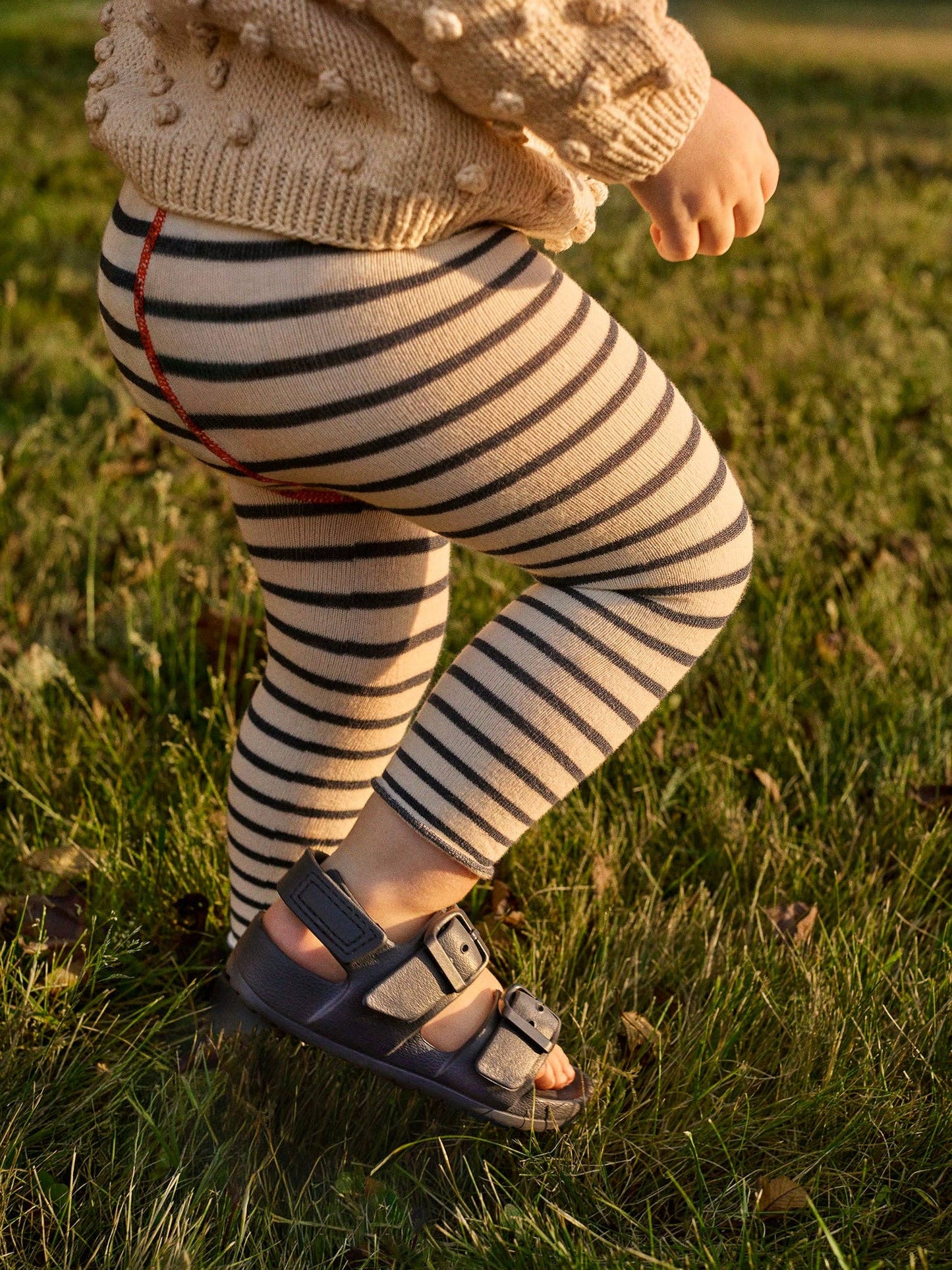 Child wearing striped leggings and a beige sweater standing on grass