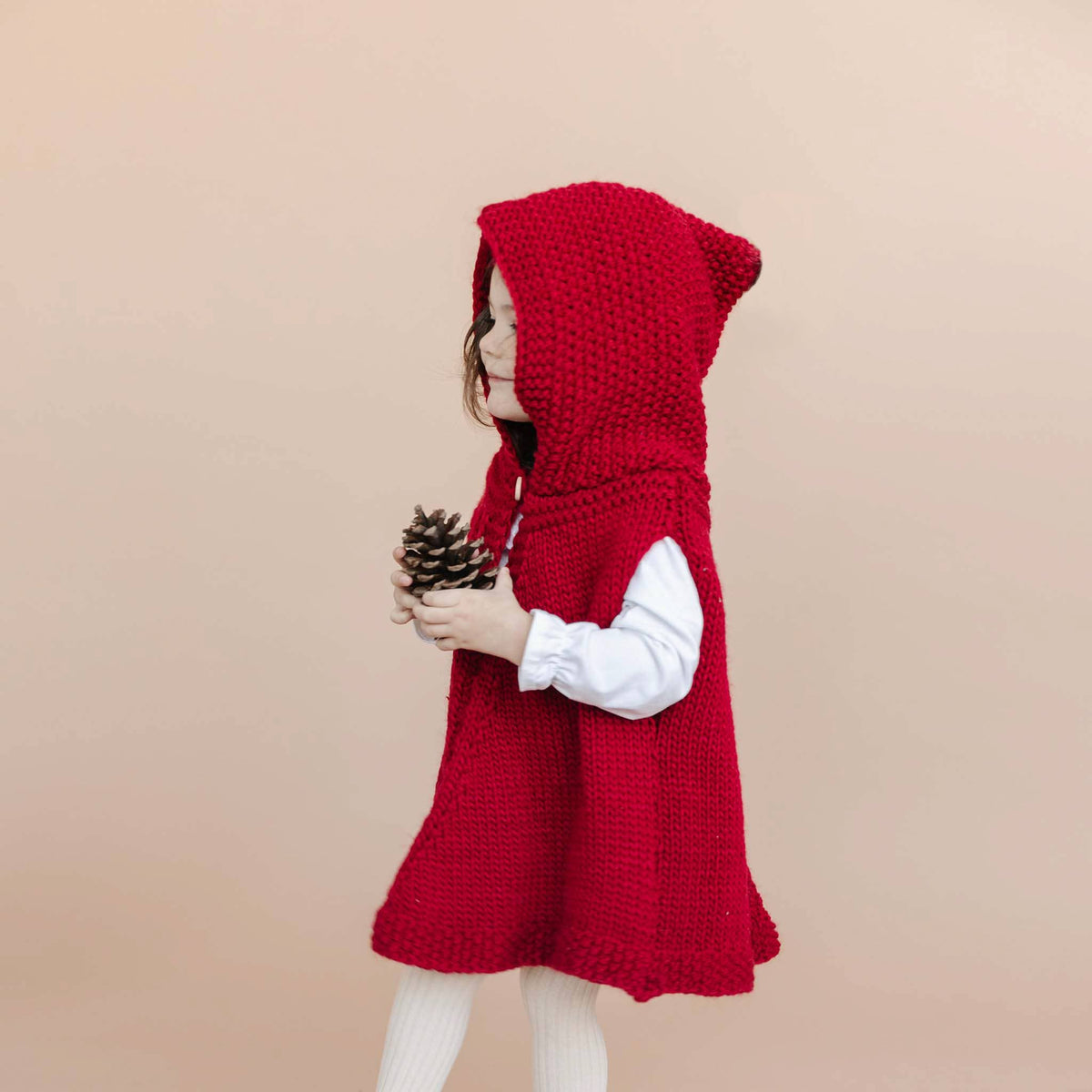A young child wearing a bright red chunky-knit hooded dress over a white long-sleeve shirt and white tights, holding a pinecone against a solid beige background.