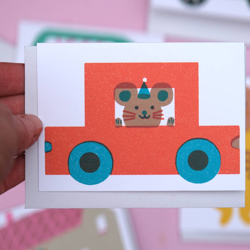 Card with a cartoon mouse in a red car held by a hand against a pink background