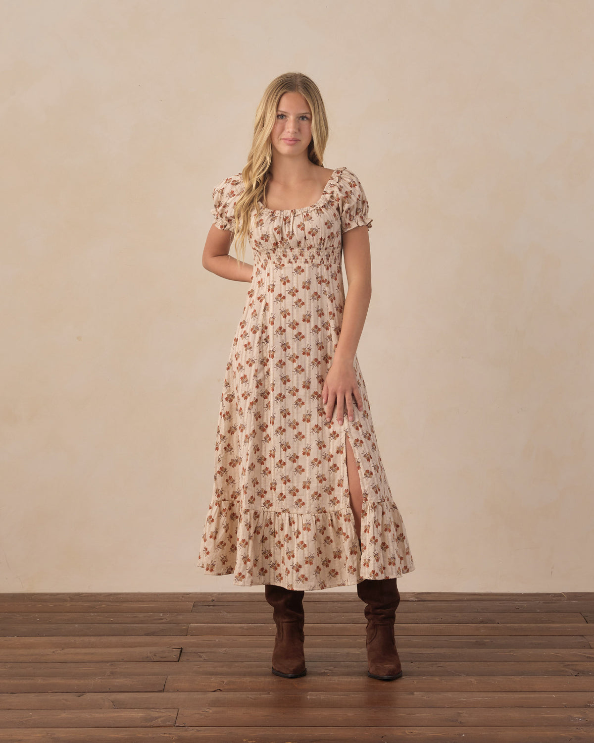 Teen wearing long floral dress and brown boots, standing in front of a beige wall on a wooden floor