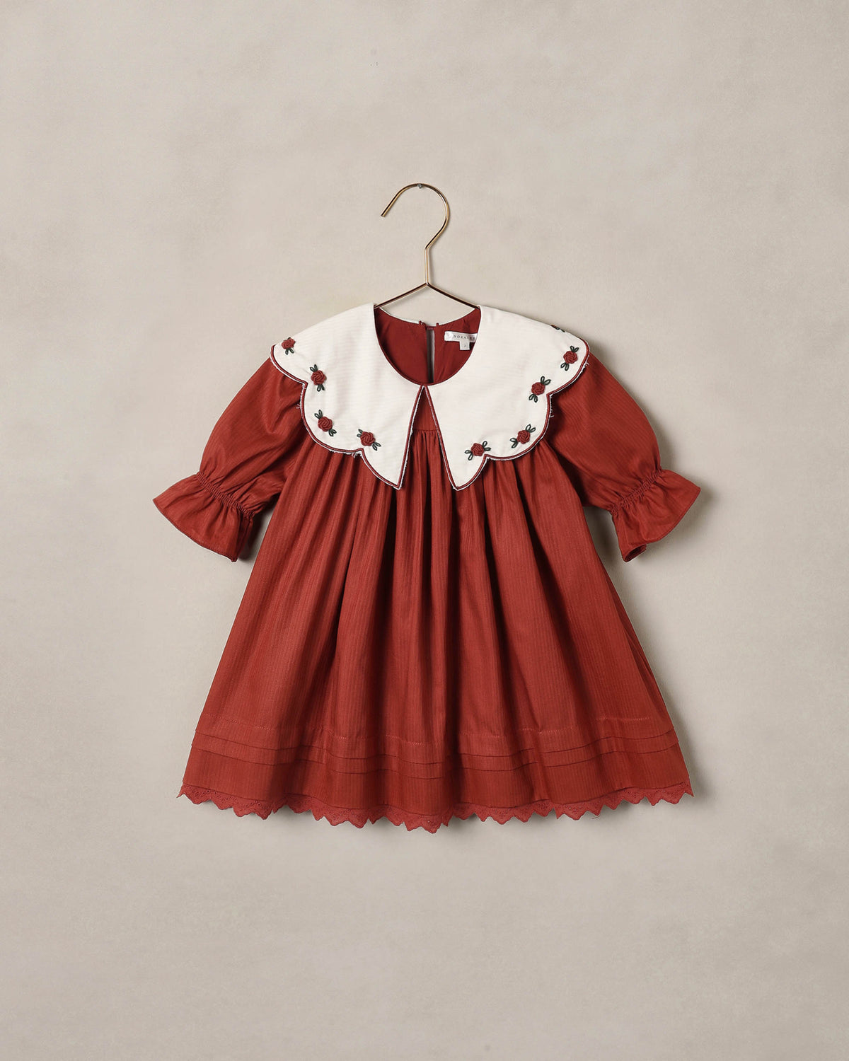 Ruby red children's dress with large white scalloped collar and pleated skirt, shown on a beige backdrop